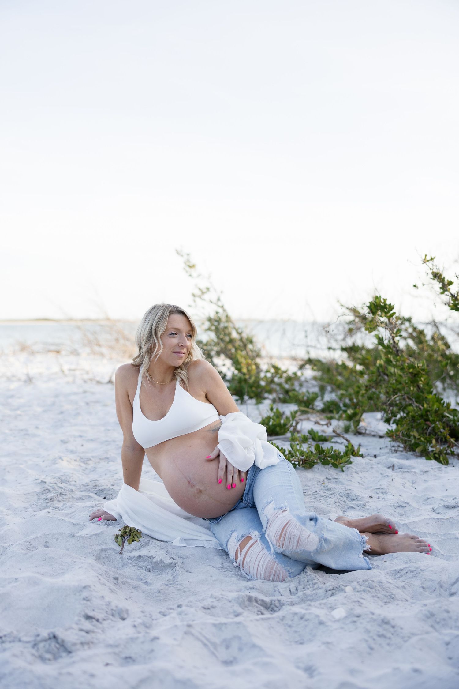 Smiling pregnant woman in white crop top and ripped jeans sitting on white sand beach.