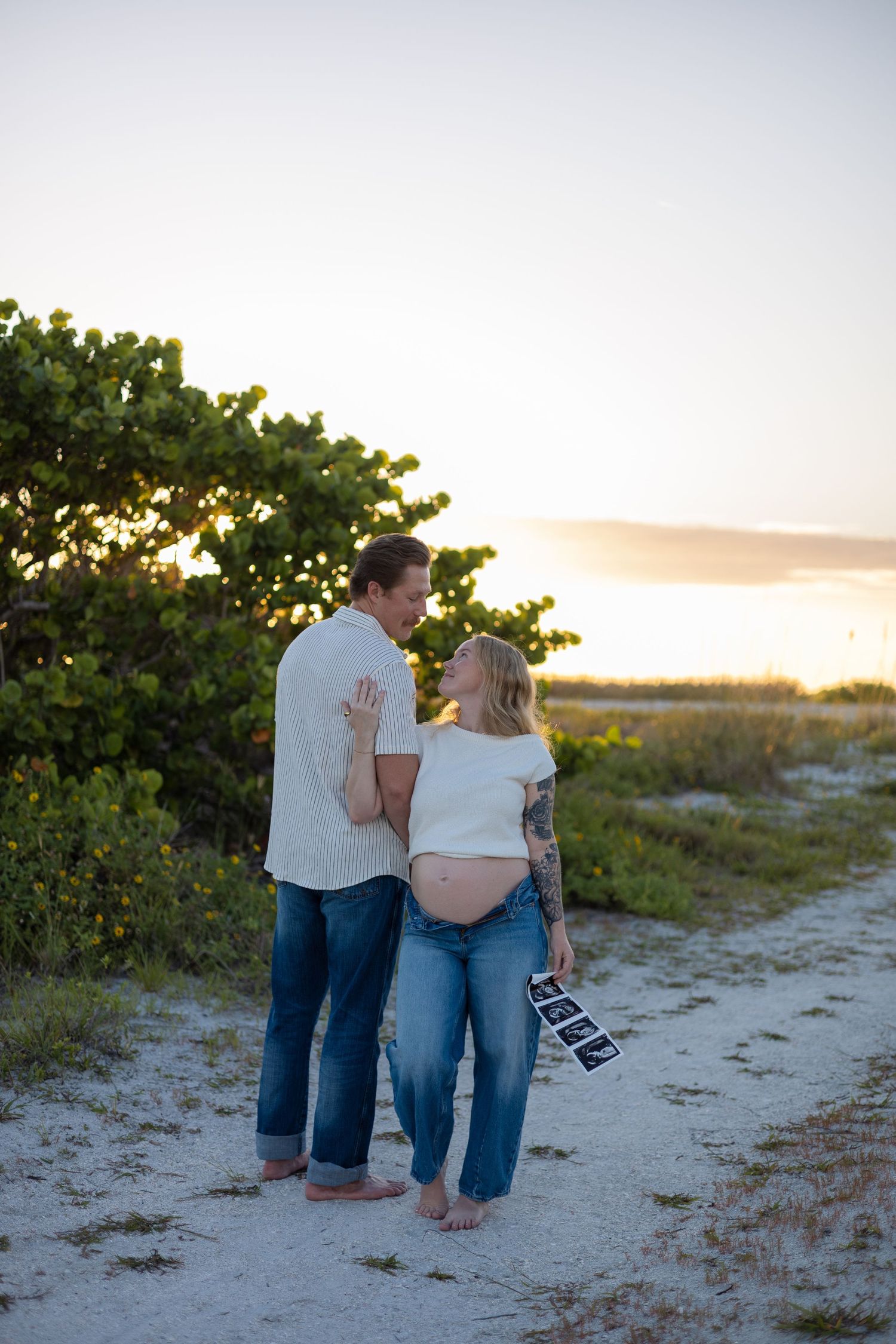 Expecting couple holding ultrasound photos while sharing a kiss on sandy beach at sunset.