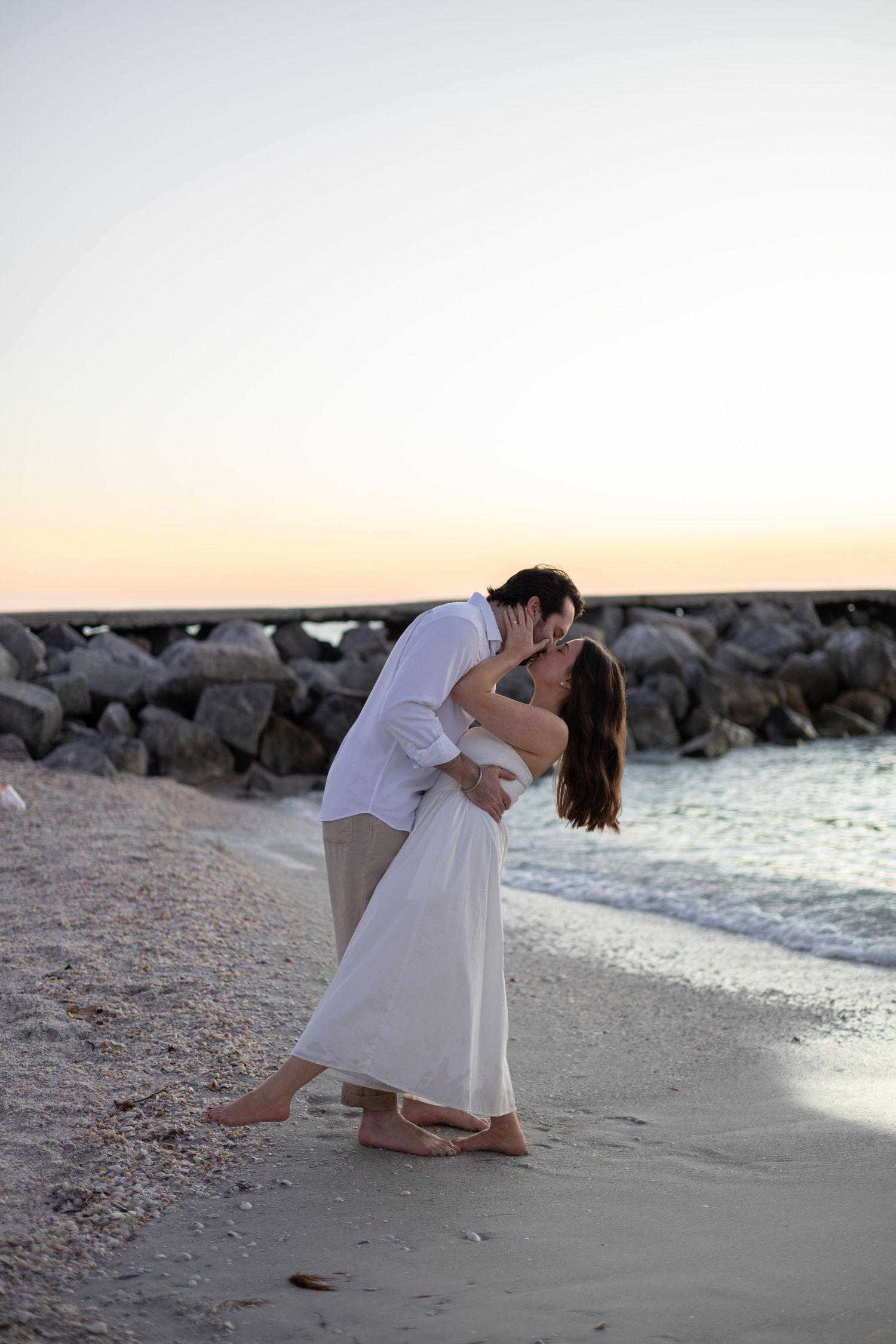Couple sharing a passionate kiss on a sandy beach at sunset near a rocky jetty.
