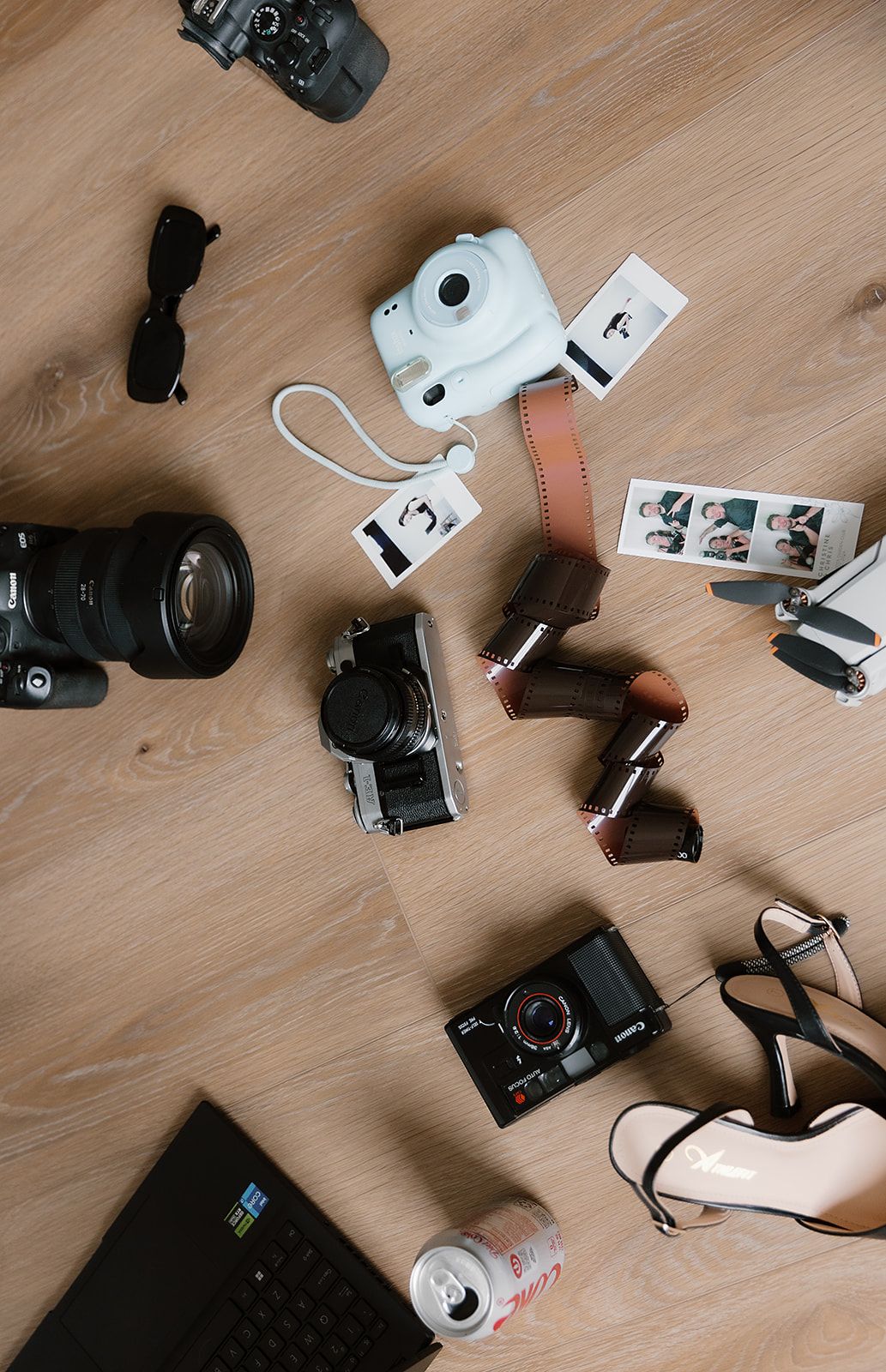 Flat lay of photography equipment including cameras, sunglasses, heels, film, and a laptop on wood floor.
