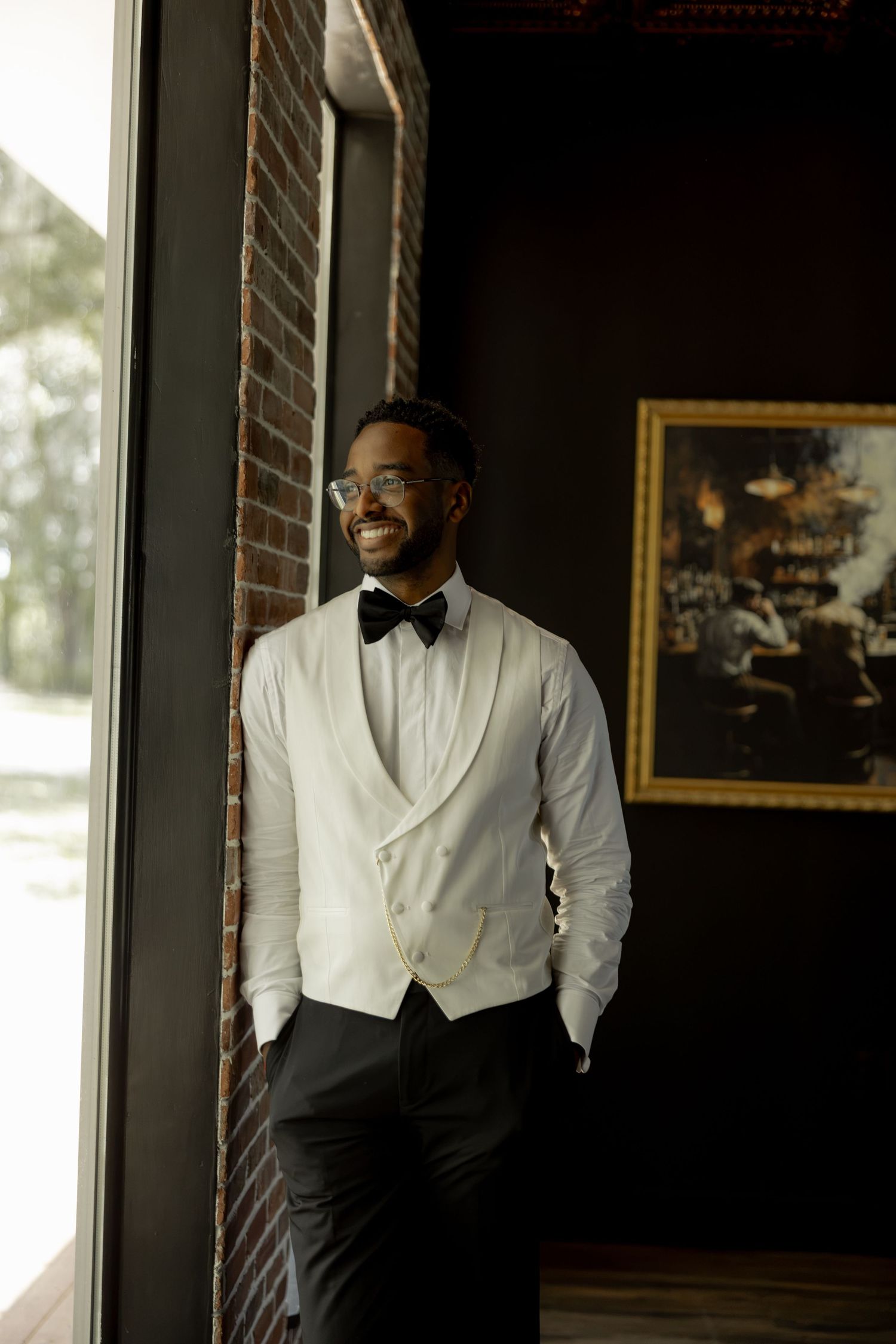 Smiling groom in white vest and black bow tie stands by window in elegant dark-walled venue.