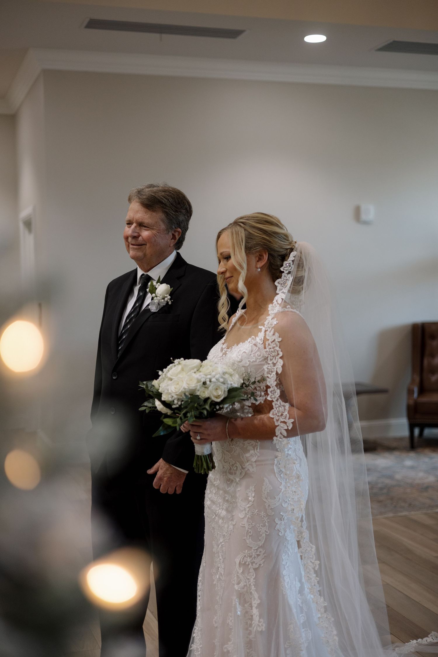A bride in lace gown holding white bouquet walks with her father toward the ceremony.