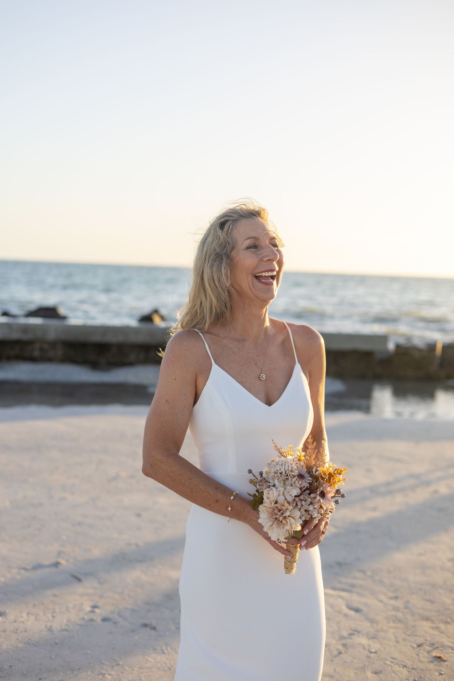 Radiant bride in white slip dress holding dried flower bouquet laughing on sandy beach at golden sunset.