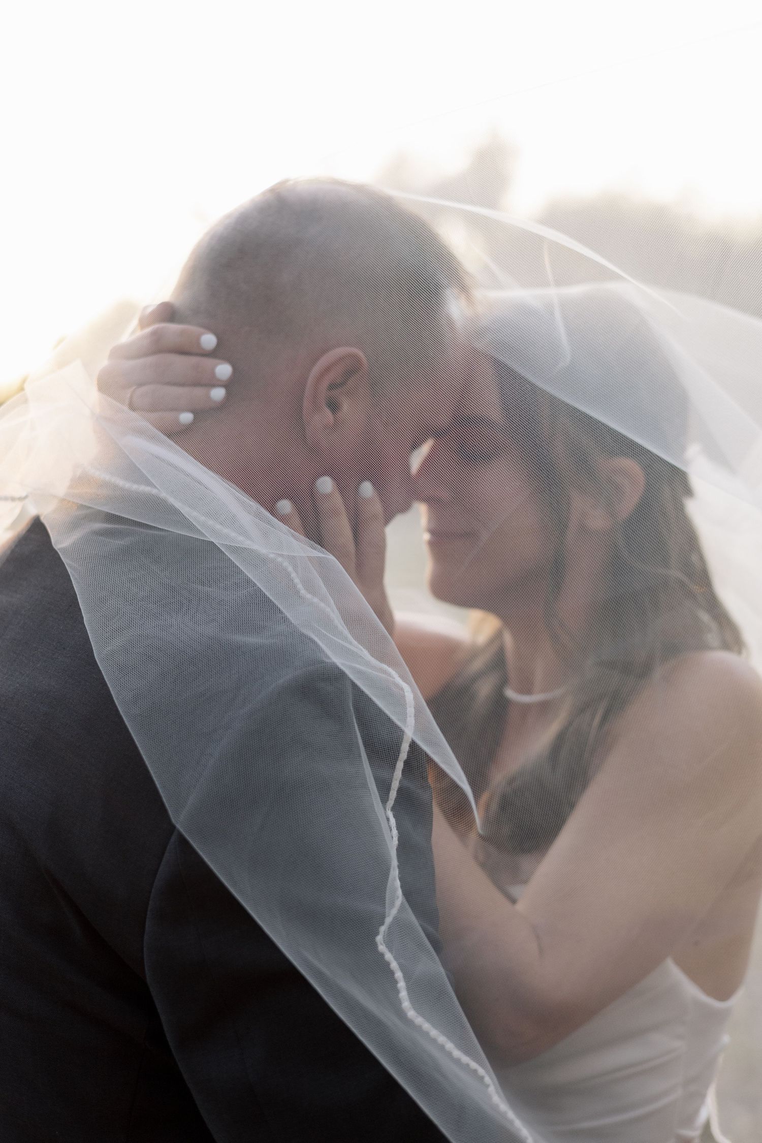 Bride and groom sharing an intimate forehead touch under a flowing veil in soft backlight.