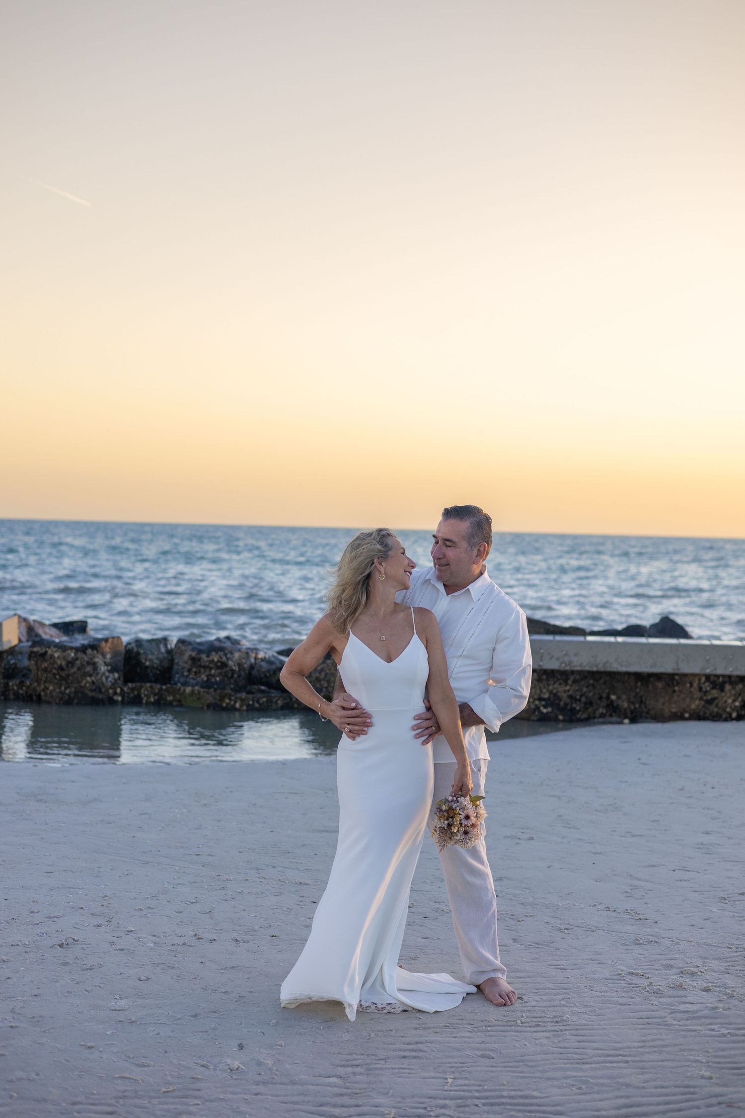 Bride and groom in white wedding attire gazing at each other on beach during golden sunset.
