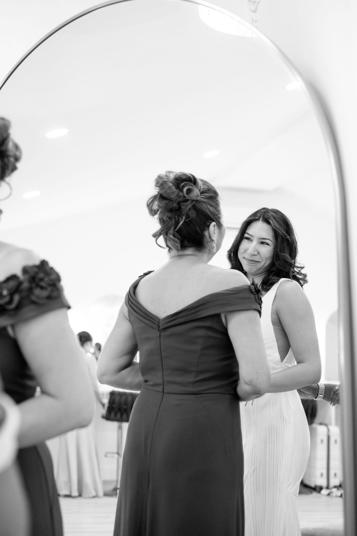 Black and white photo of bride smiling at woman in dark gown reflected in arched mirror.