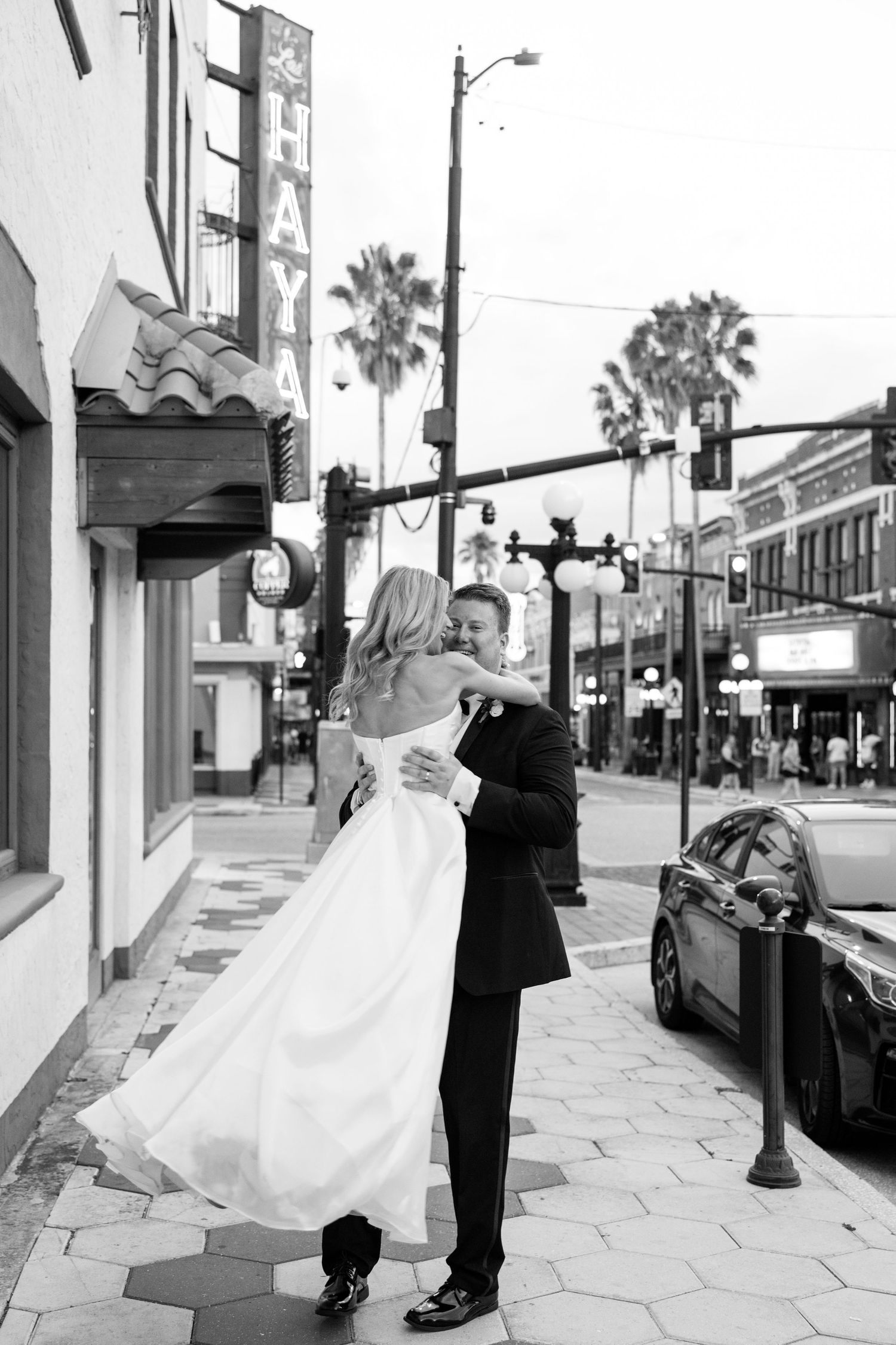 Black and white photo of newlyweds embracing on Ybor City sidewalk with palm trees backdrop.