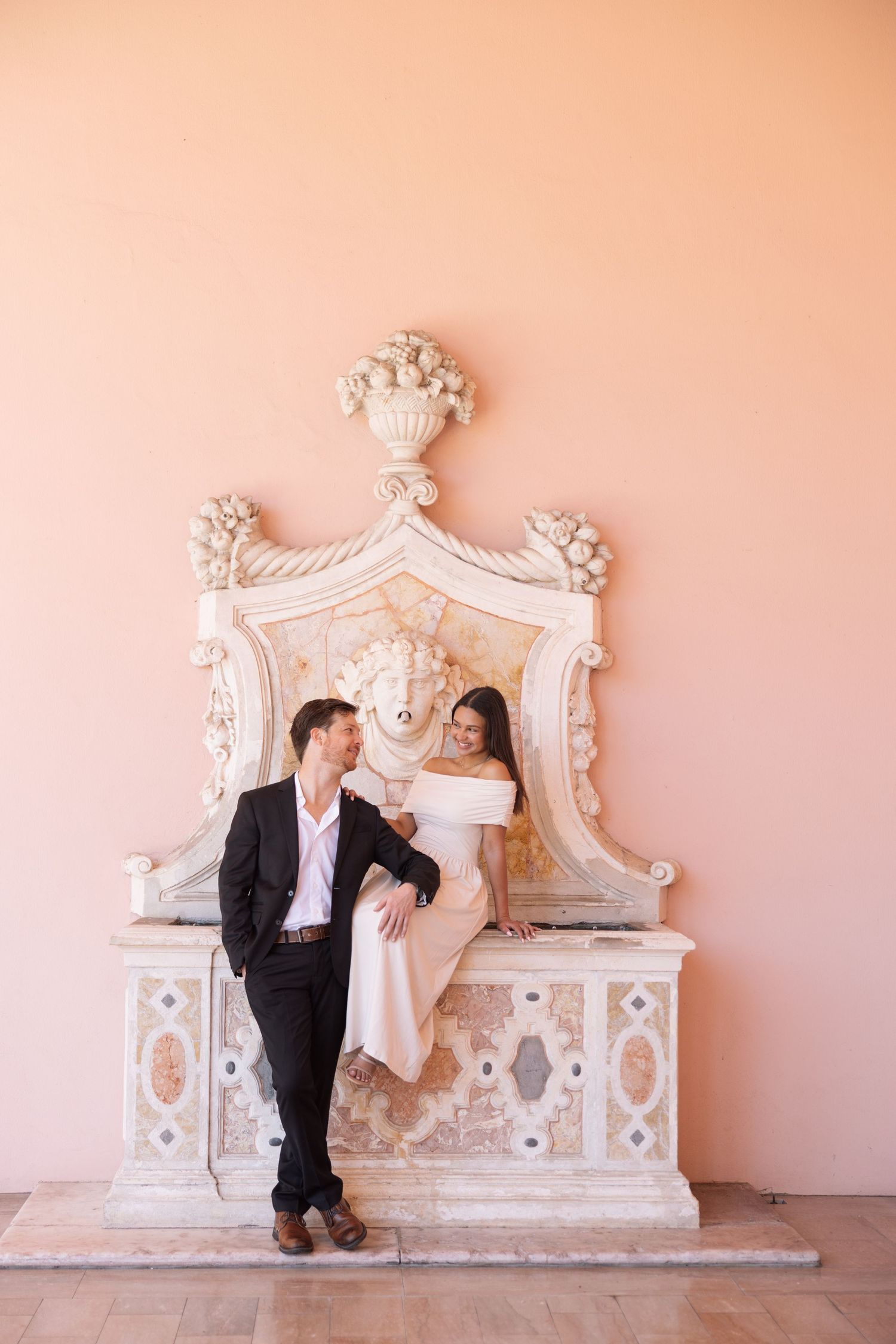 Elegant couple posing beside ornate antique fountain against soft pink wall in historic venue.