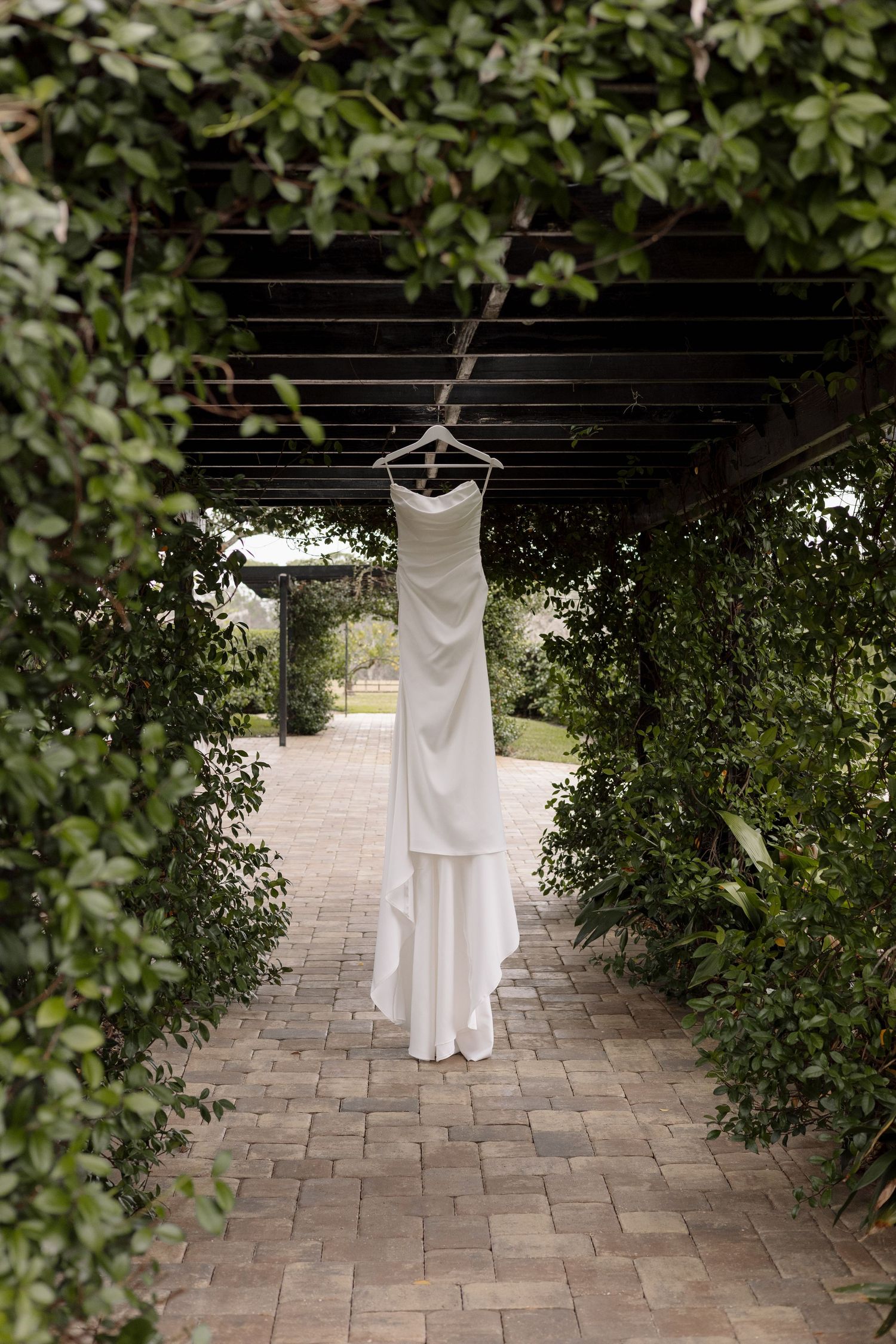 White wedding dress hanging on a hanger under a vine-covered pergola in a lush garden walkway.