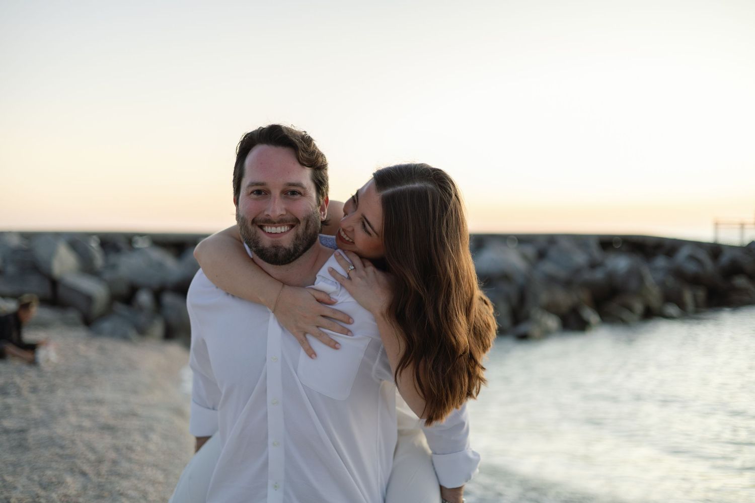 Happy couple at the beach, woman hugging man from behind and kissing his cheek at sunset.