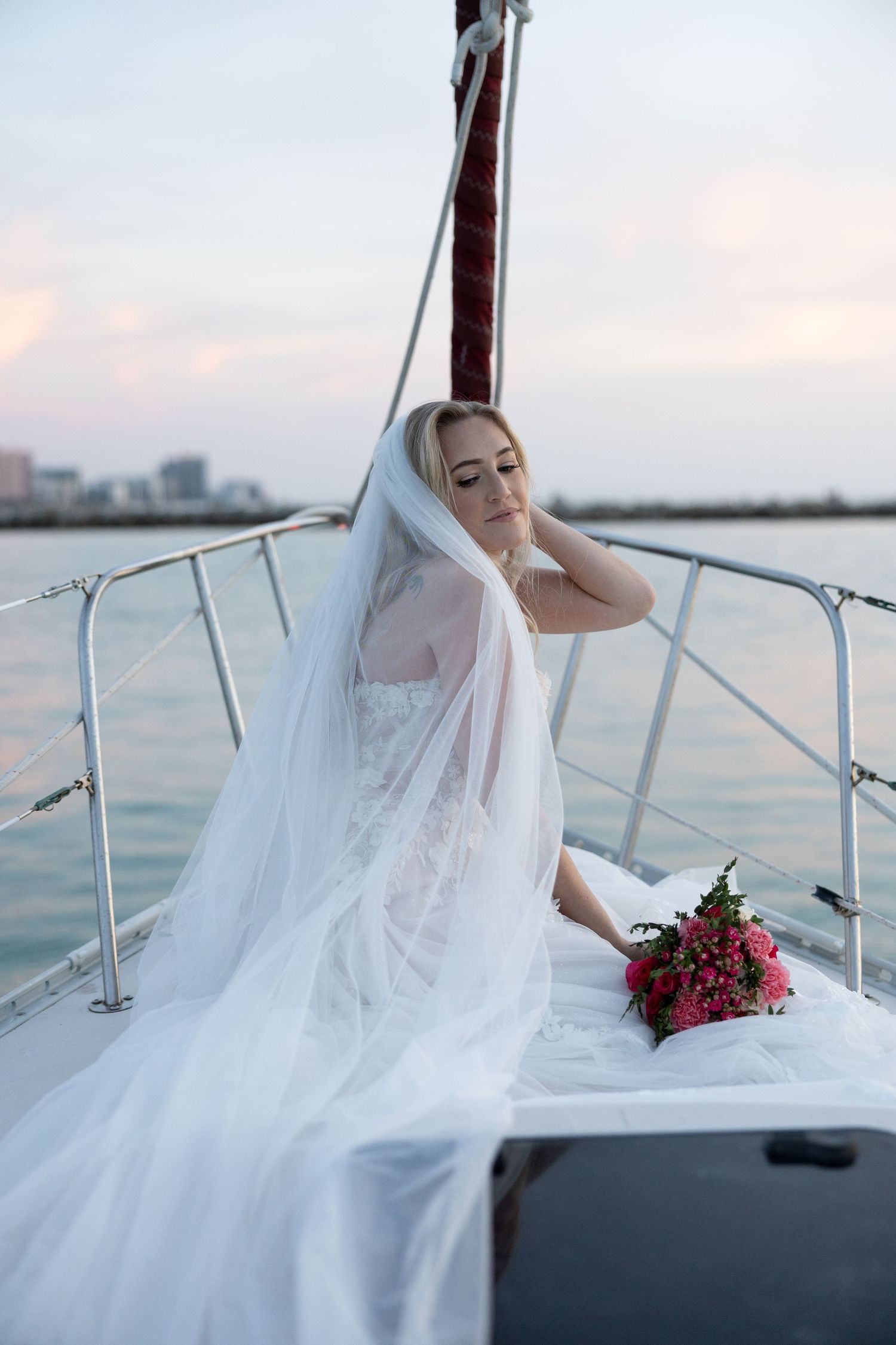 Bride in white gown and veil sits on sailboat deck holding red bouquet with city skyline at dusk.