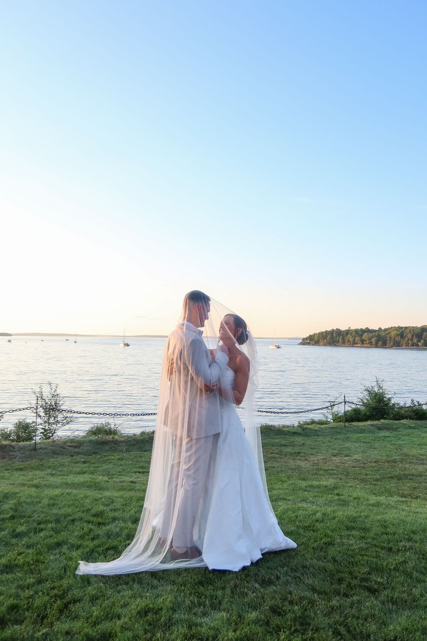 Bride and groom embracing at sunset by the water, bride wearing long veil and white gown on green lawn.