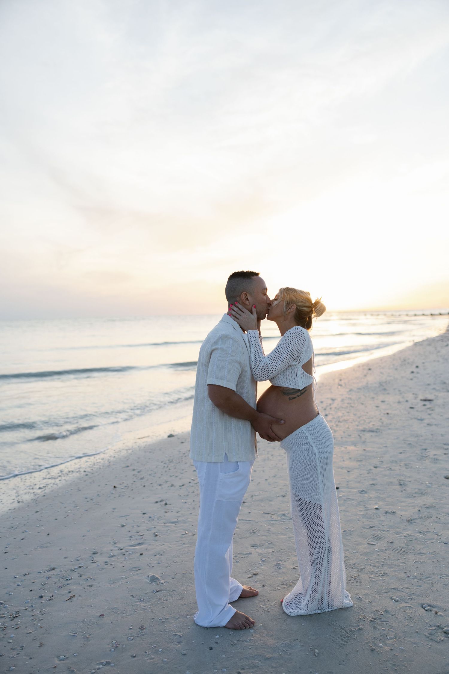 Pregnant couple sharing a kiss on the beach at sunset with ocean in background.