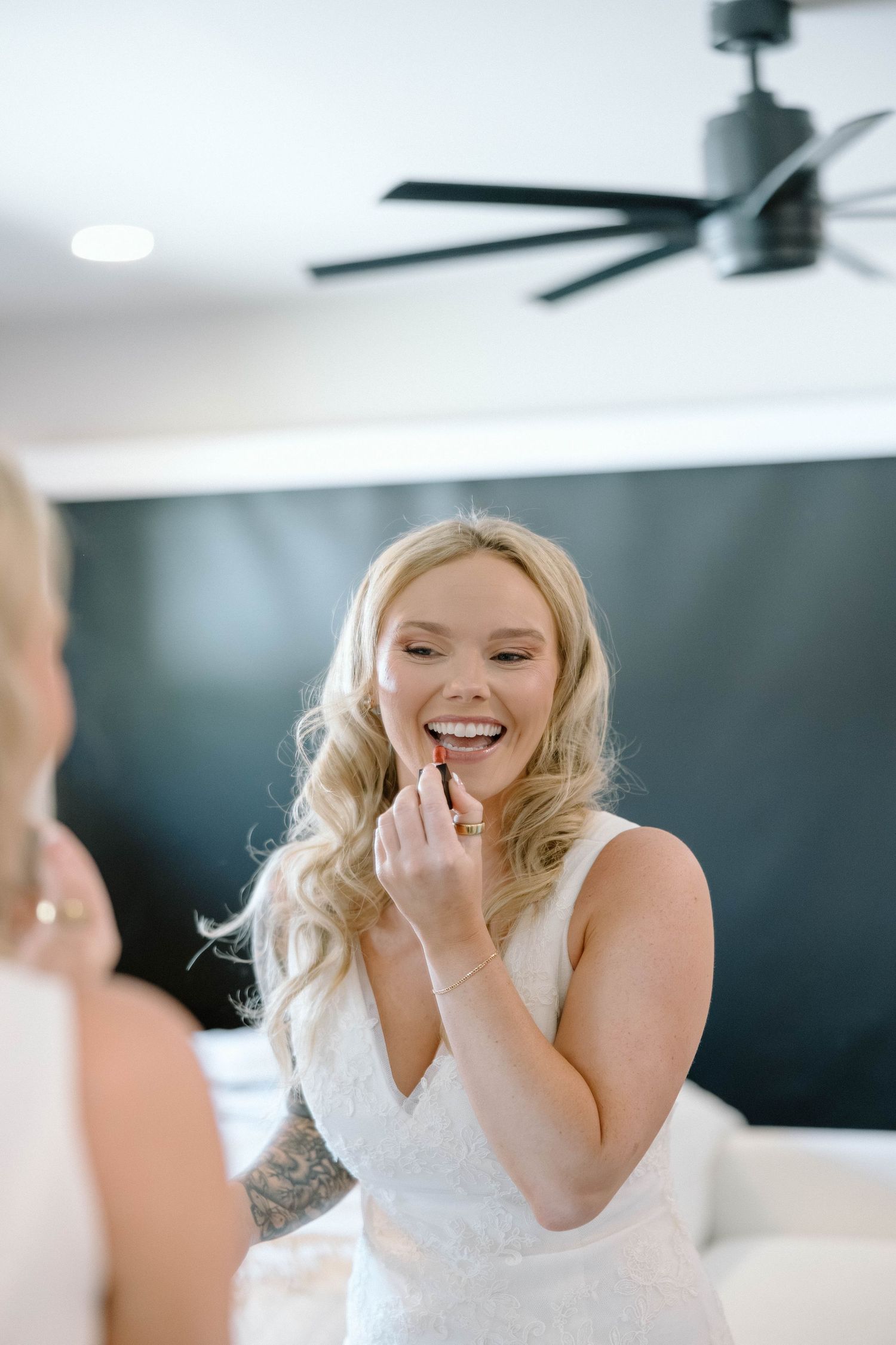 Smiling blonde bride applying lipstick in mirror while wearing white wedding dress during bridal preparations.