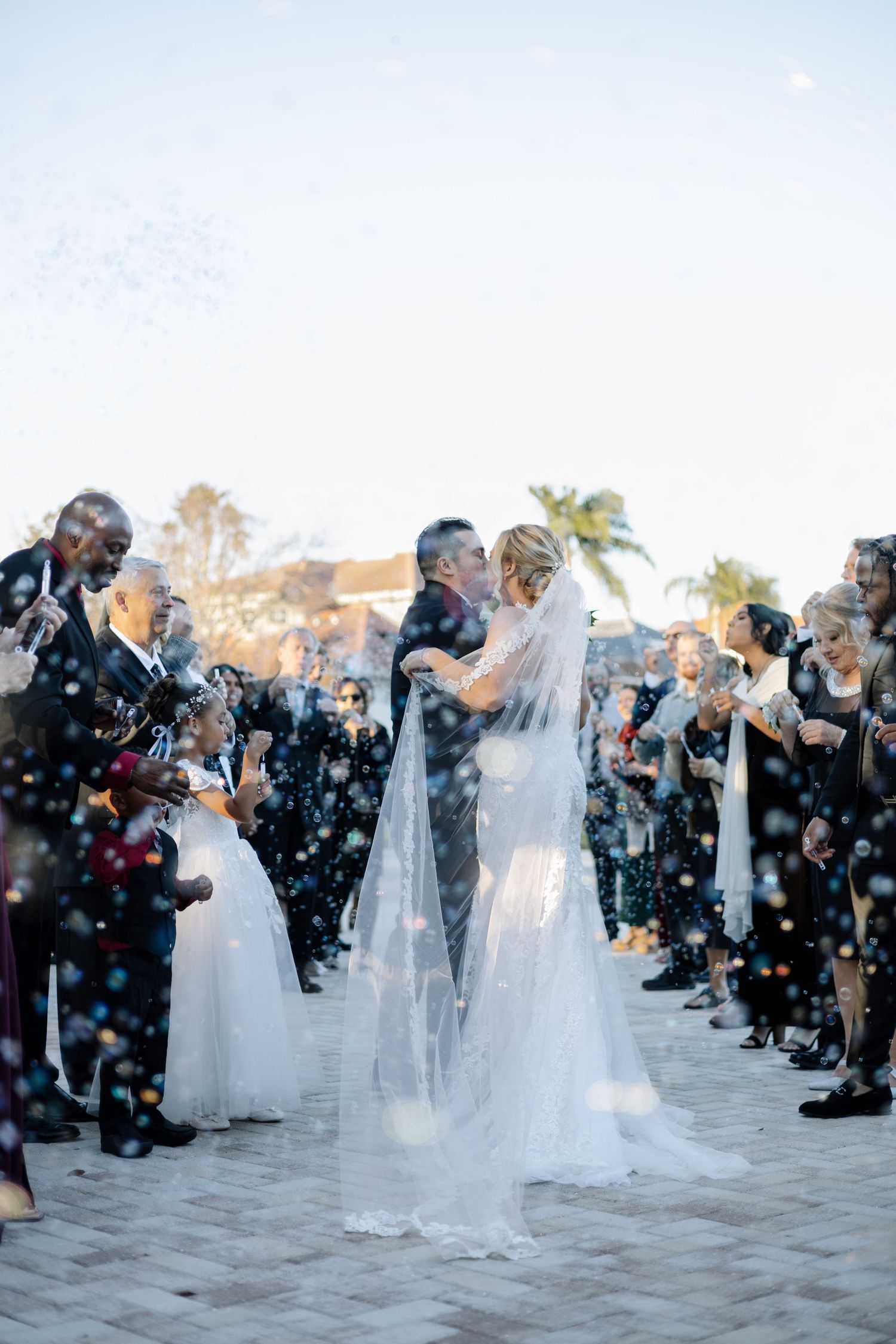 Bride and groom kiss during joyful outdoor bubble exit surrounded by cheering wedding guests.