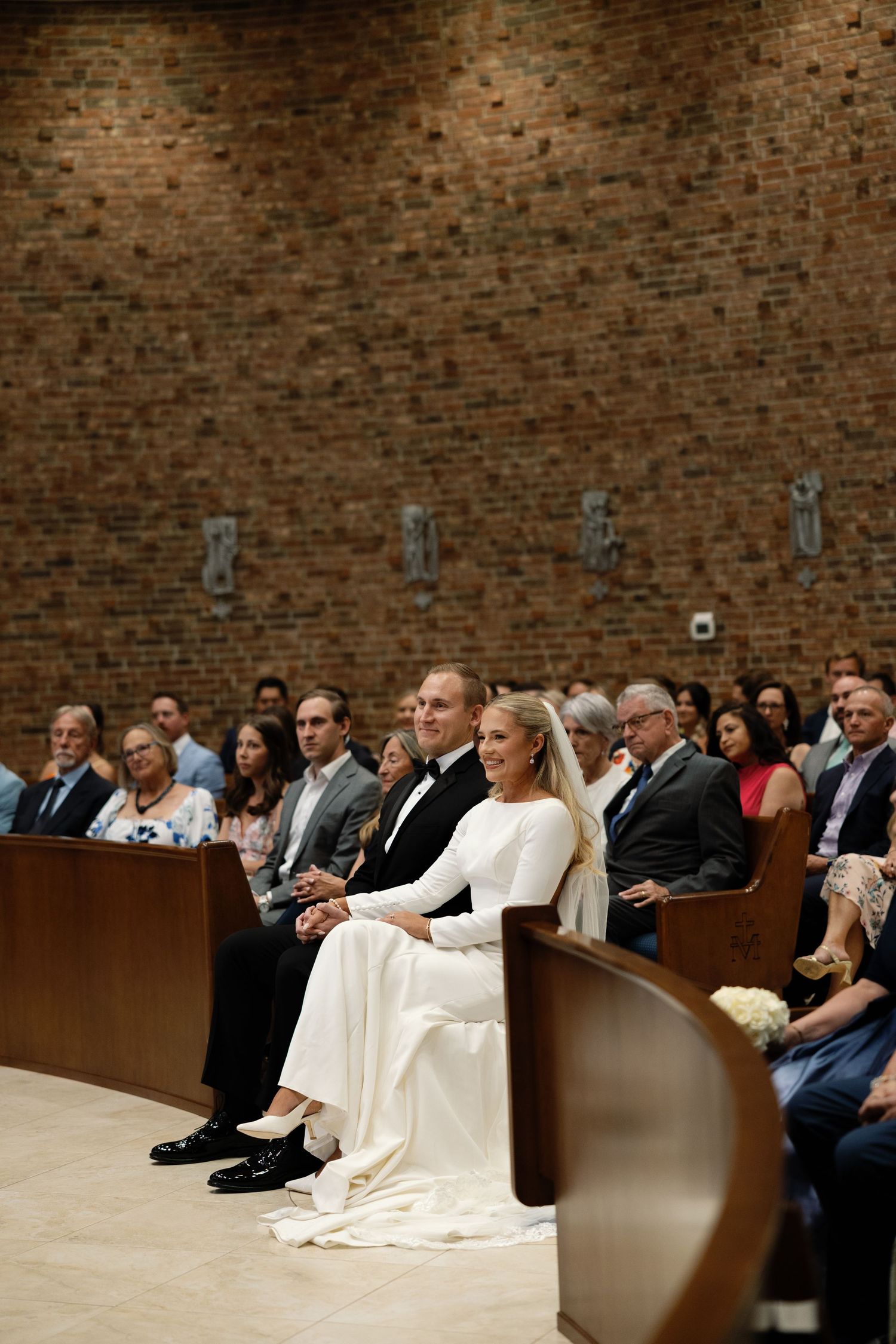 Bride and groom seated in church pews smiling during wedding ceremony with guests.