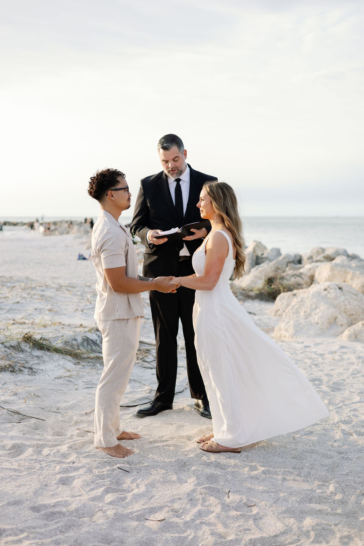 Same-sex couple exchanges vows barefoot on sandy beach at sunset with officiant reading ceremony.