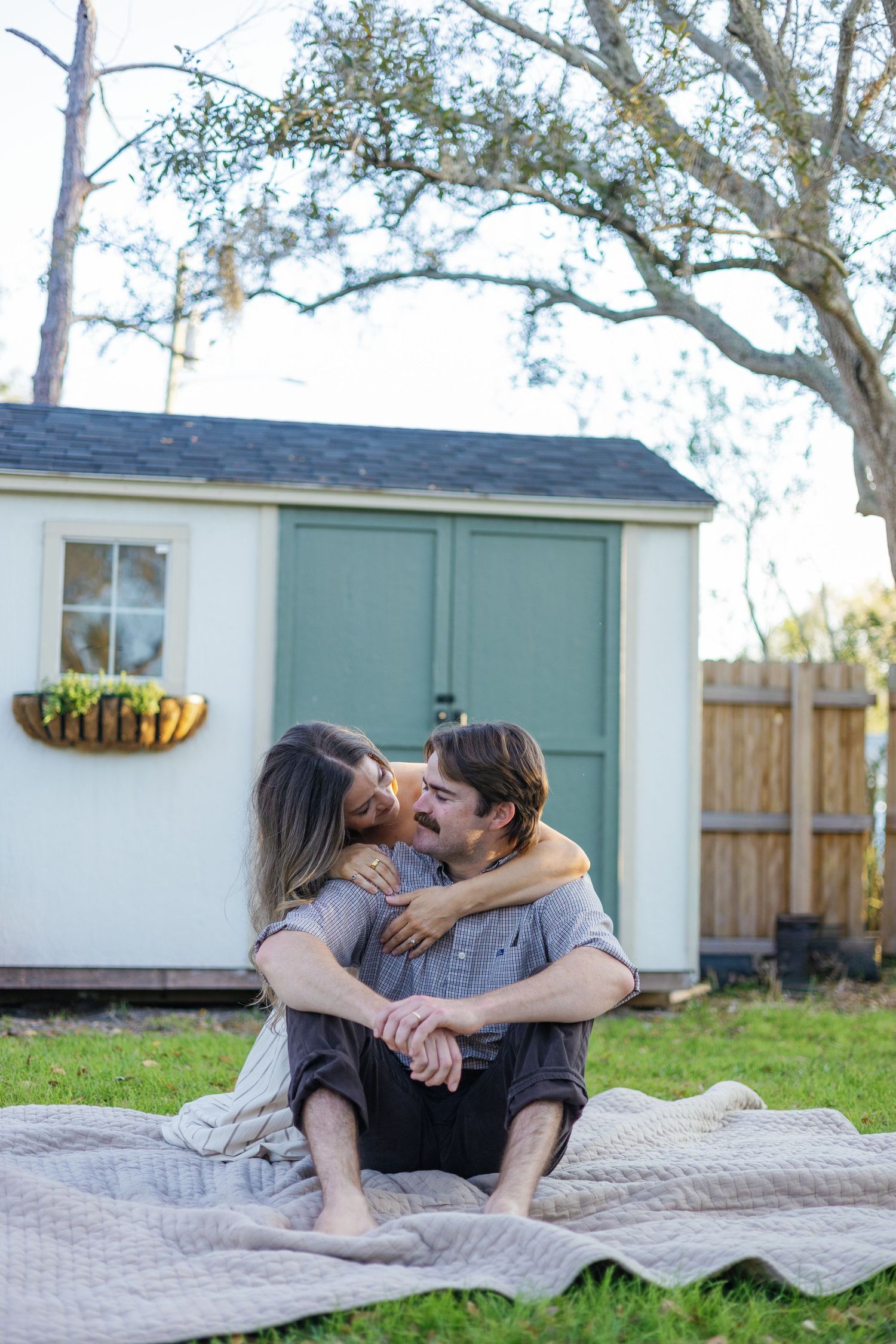 Couple sitting on a blanket in a backyard, sharing a tender kiss near a shed.
