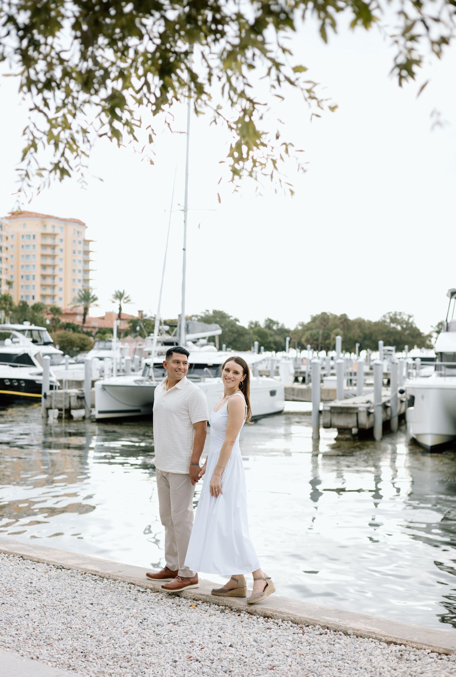 Couple posing hand-in-hand along a scenic marina waterfront with sailboats and a waterfront hotel backdrop.