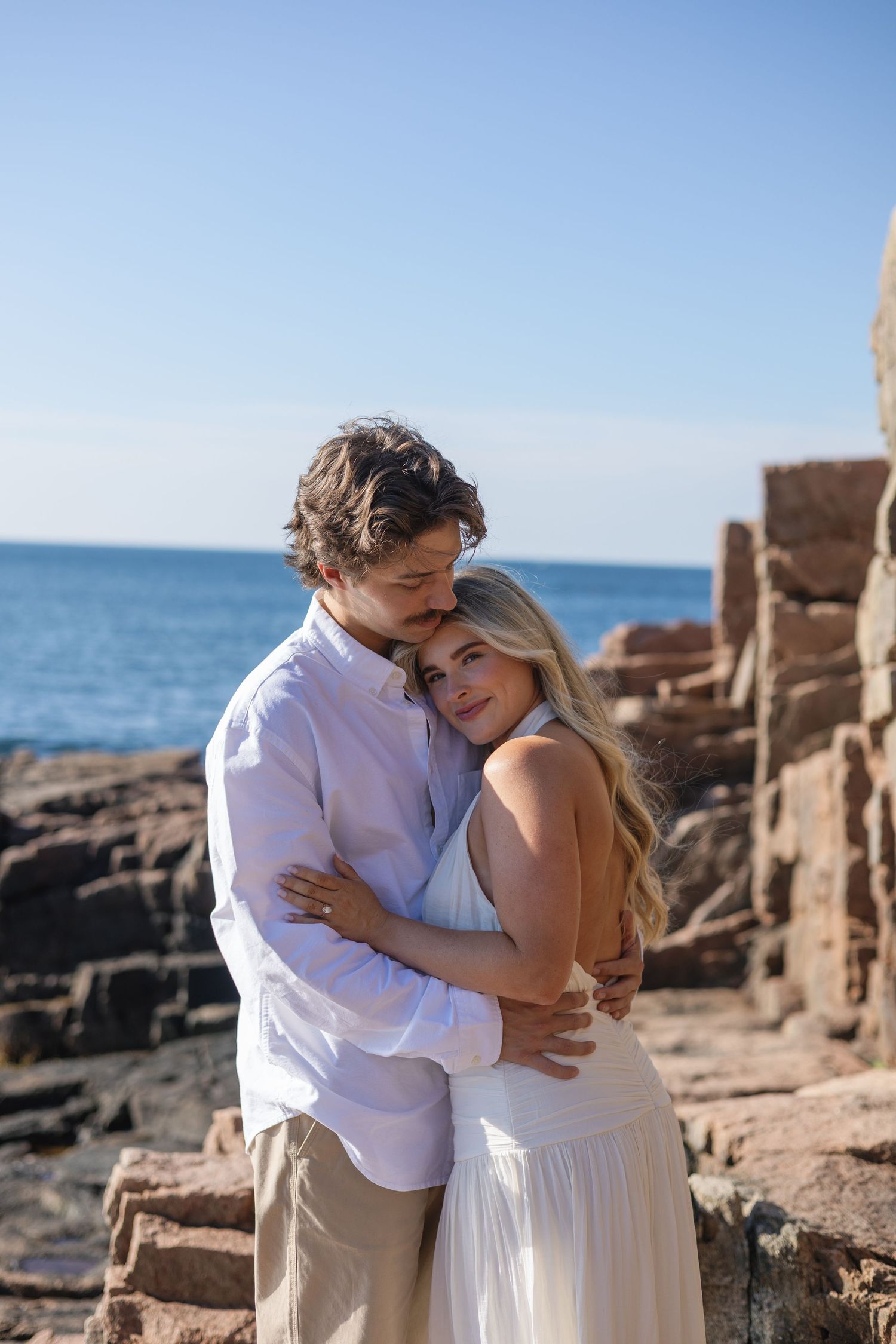 A loving couple embraces near rocky coastal ruins with the blue ocean and clear sky in the background.