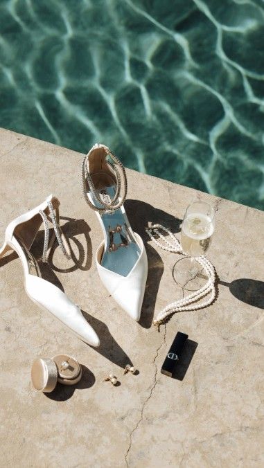White bridal heels with crystal ankle straps beside champagne glass and accessories on marble poolside.