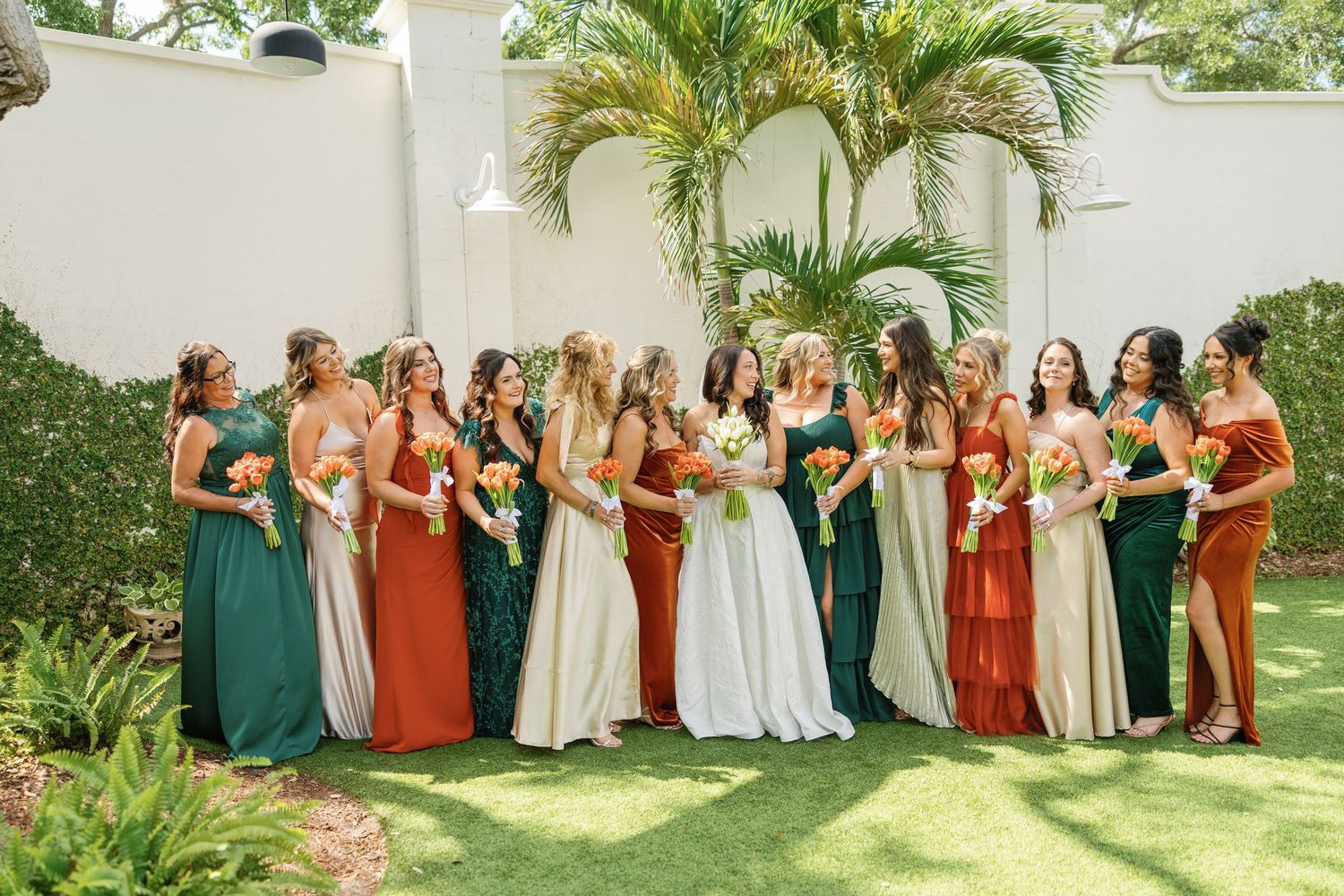 Bride poses with bridesmaids in mixed earth-tone dresses holding colorful bouquets in a tropical garden.