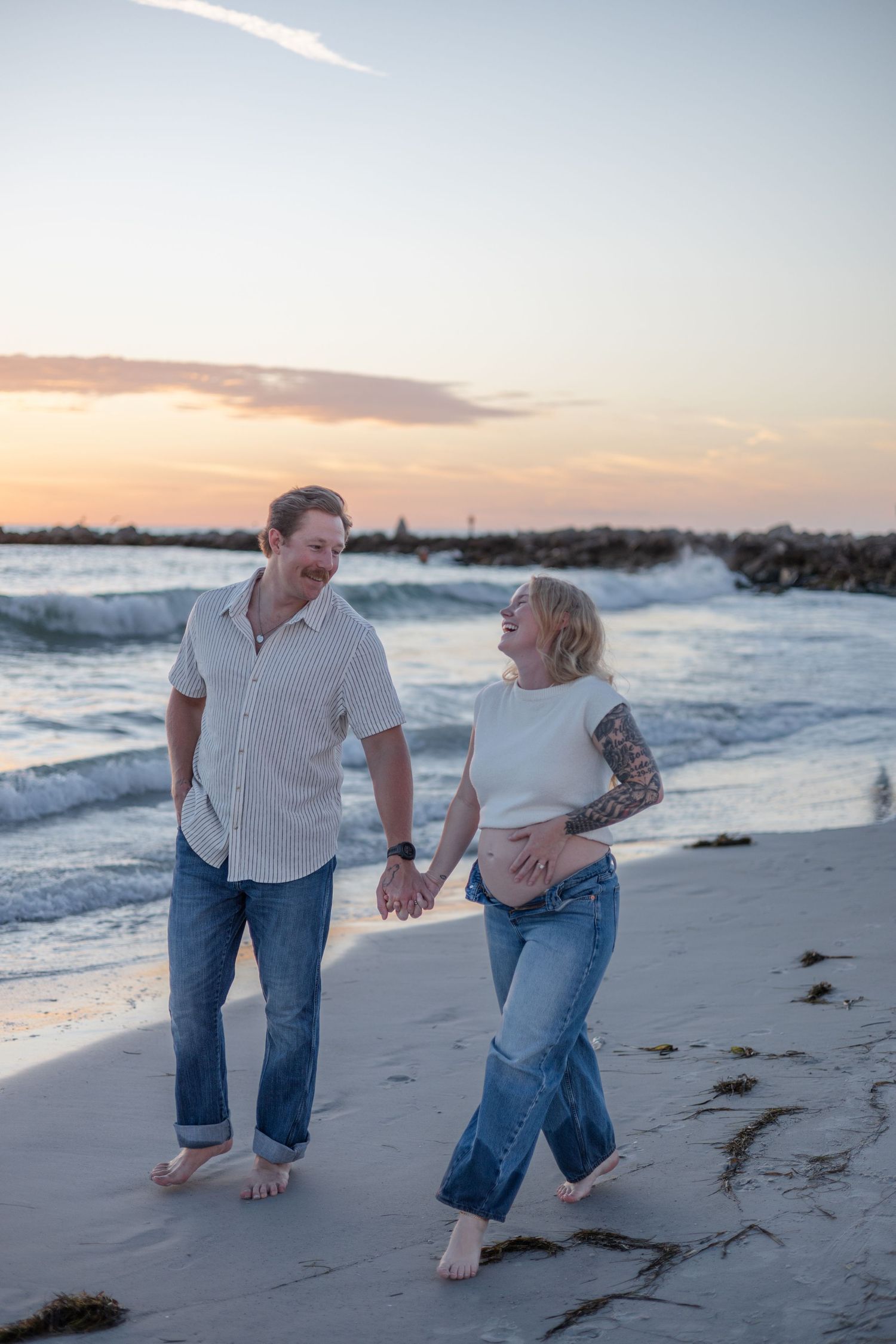 Happy expecting couple walking hand-in-hand along sandy beach at colorful sunset, smiling at each other.