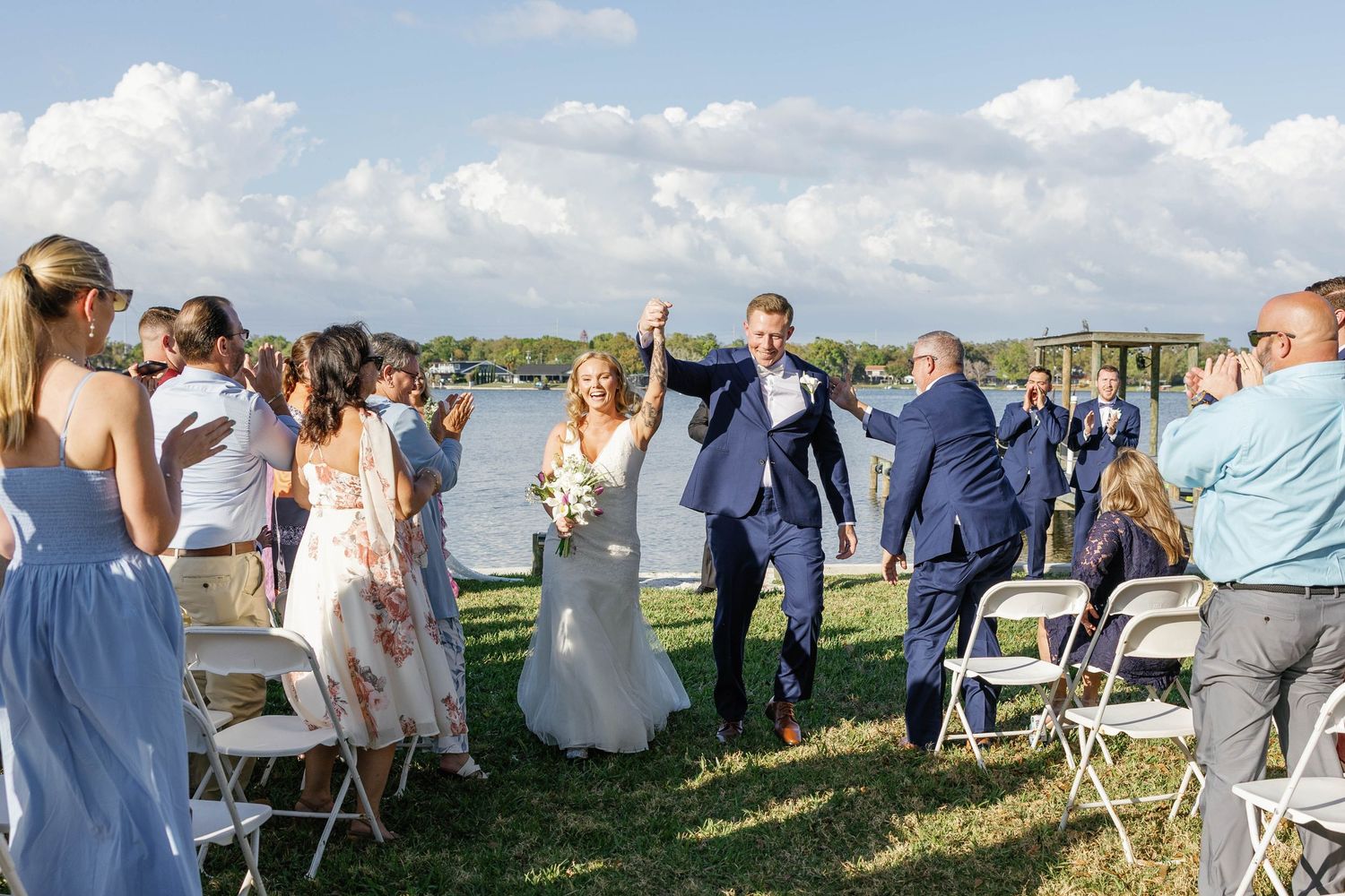 Joyful bride and groom raising hands walking back down the aisle after outdoor lakeside wedding ceremony.