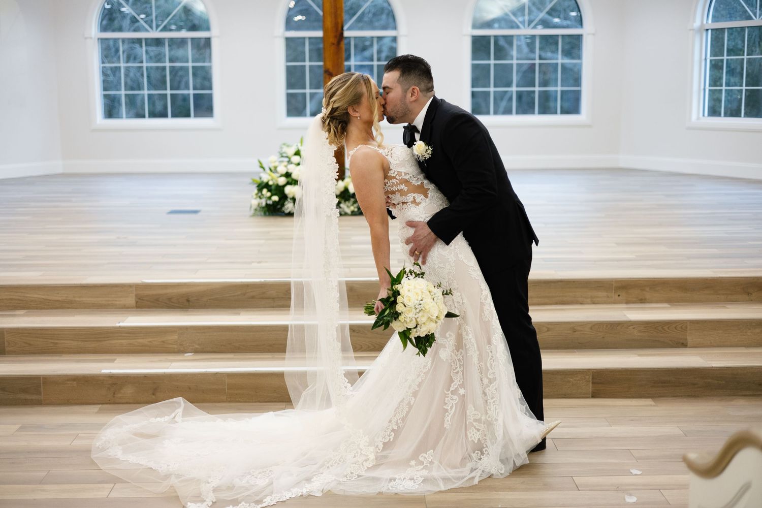 Bride and groom share romantic kiss at chapel altar with wooden cross and large windows.
