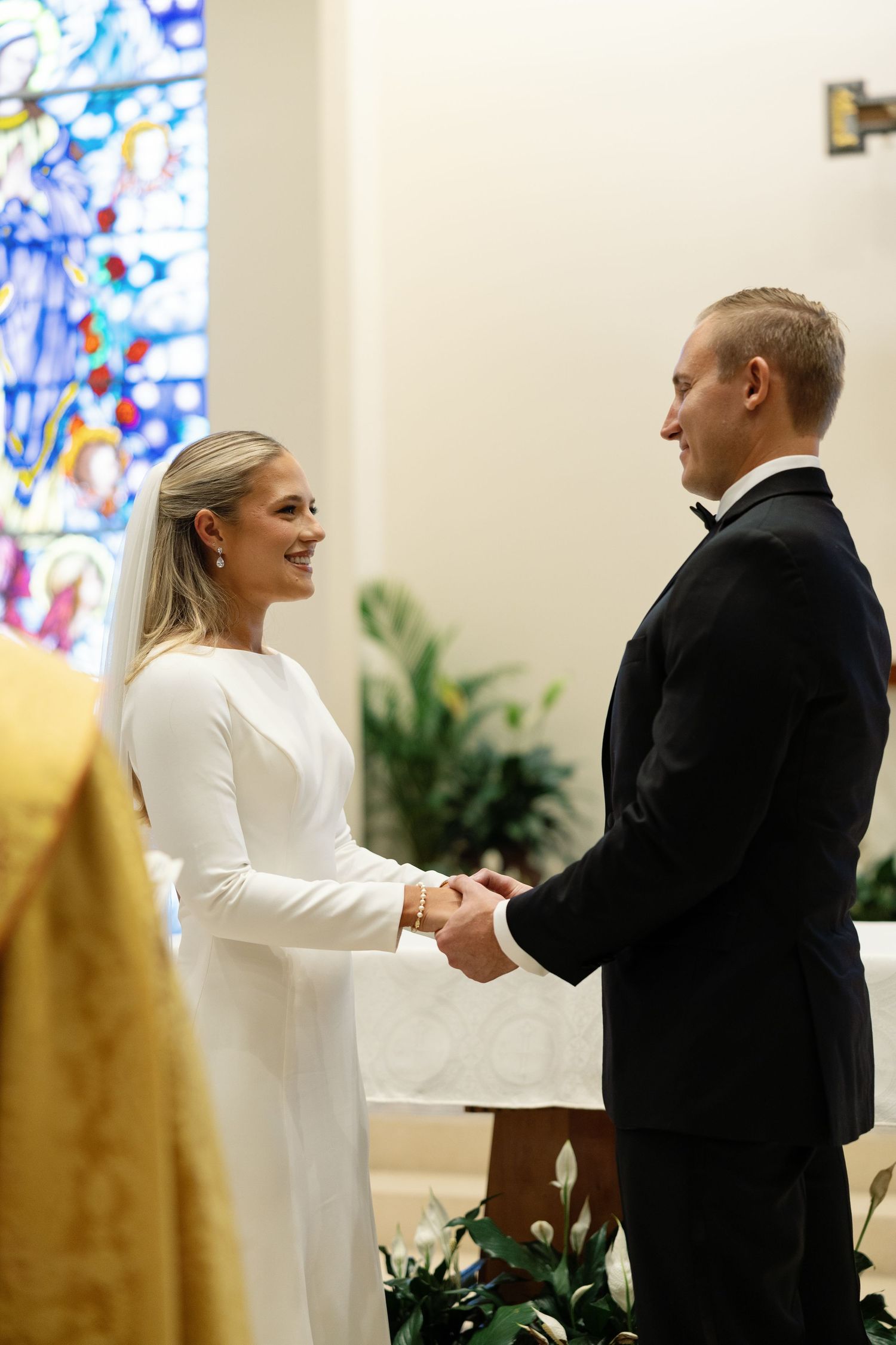 Bride and groom holding hands and exchanging vows before stained glass church window.