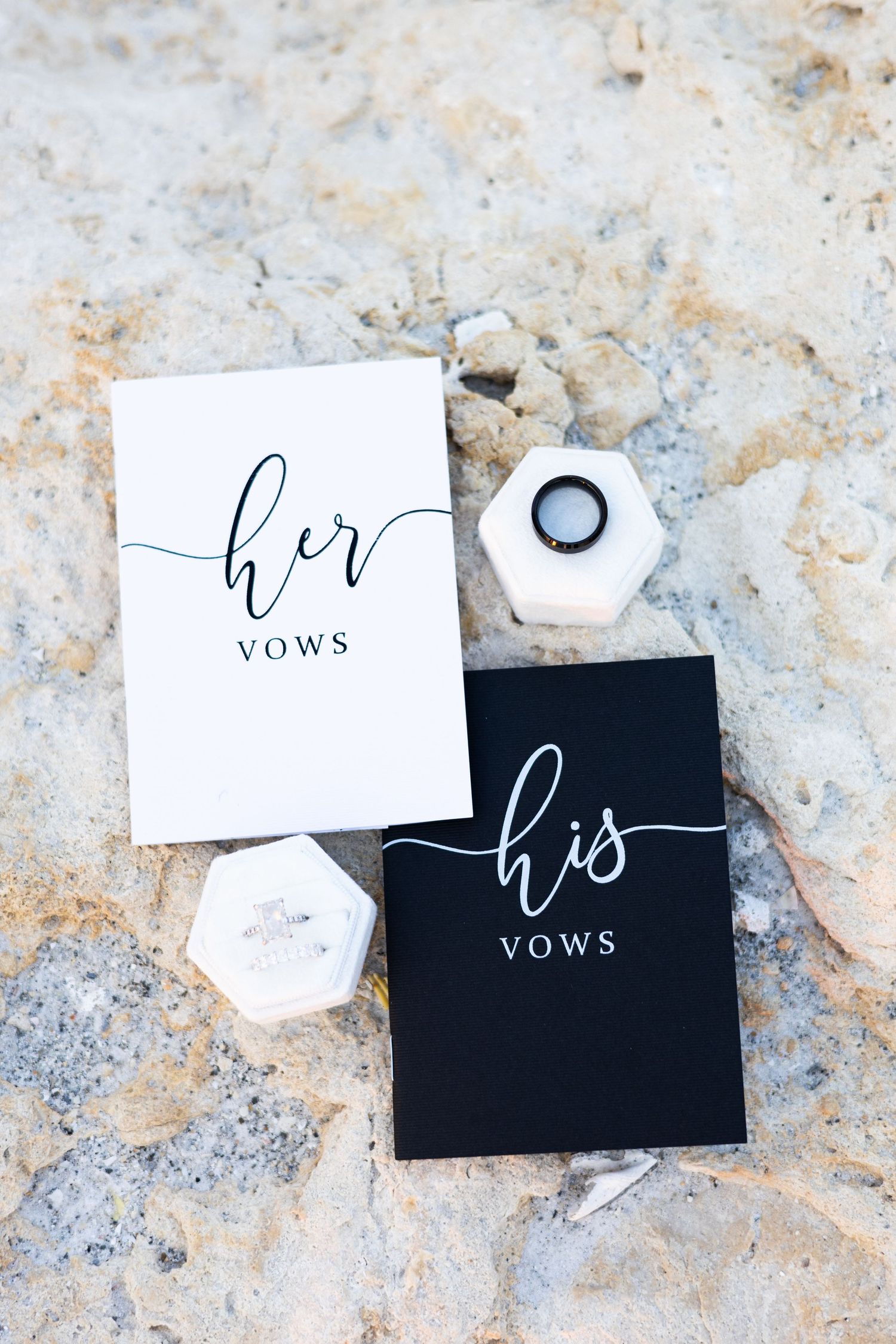 His and her vows booklets with wedding rings in white hexagon box on rocky surface.