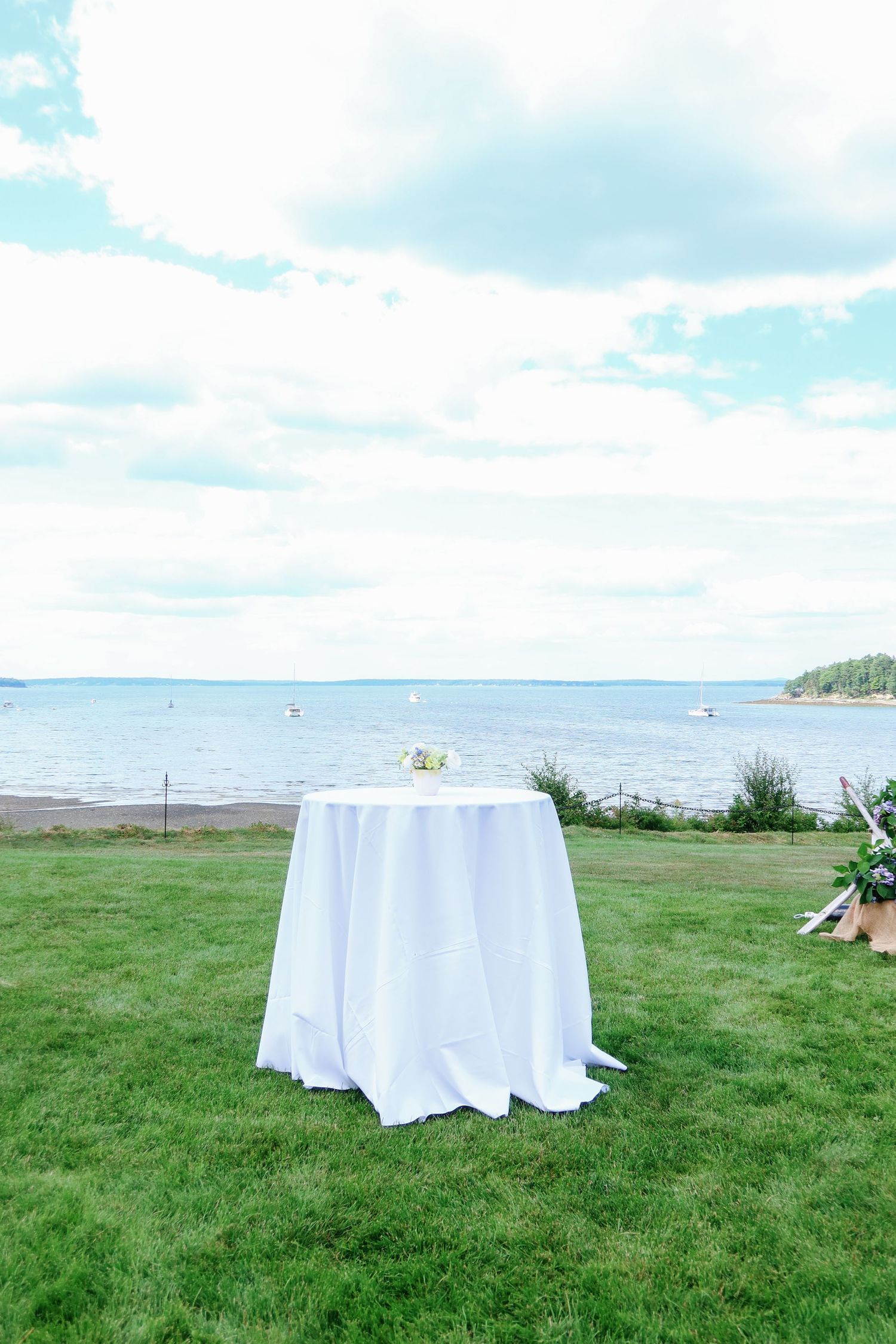 White linen cocktail table on green lawn with serene ocean view and cloudy blue sky at outdoor wedding.