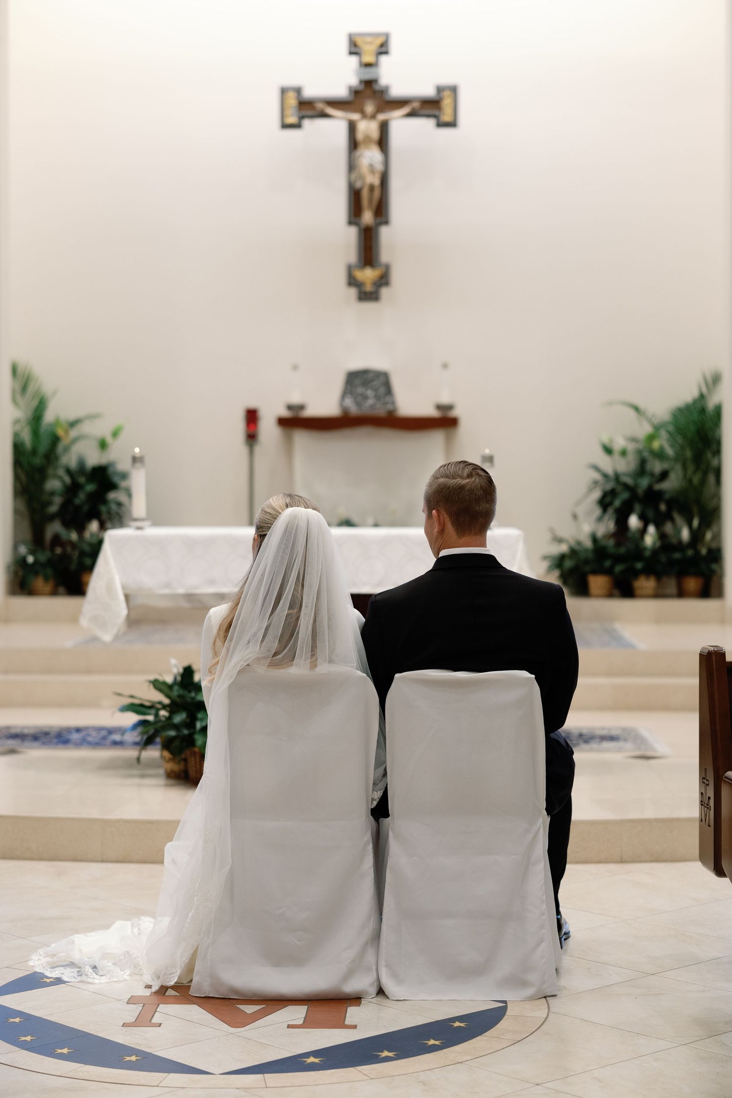 Bride and groom seated at altar before crucifix during Catholic wedding ceremony.