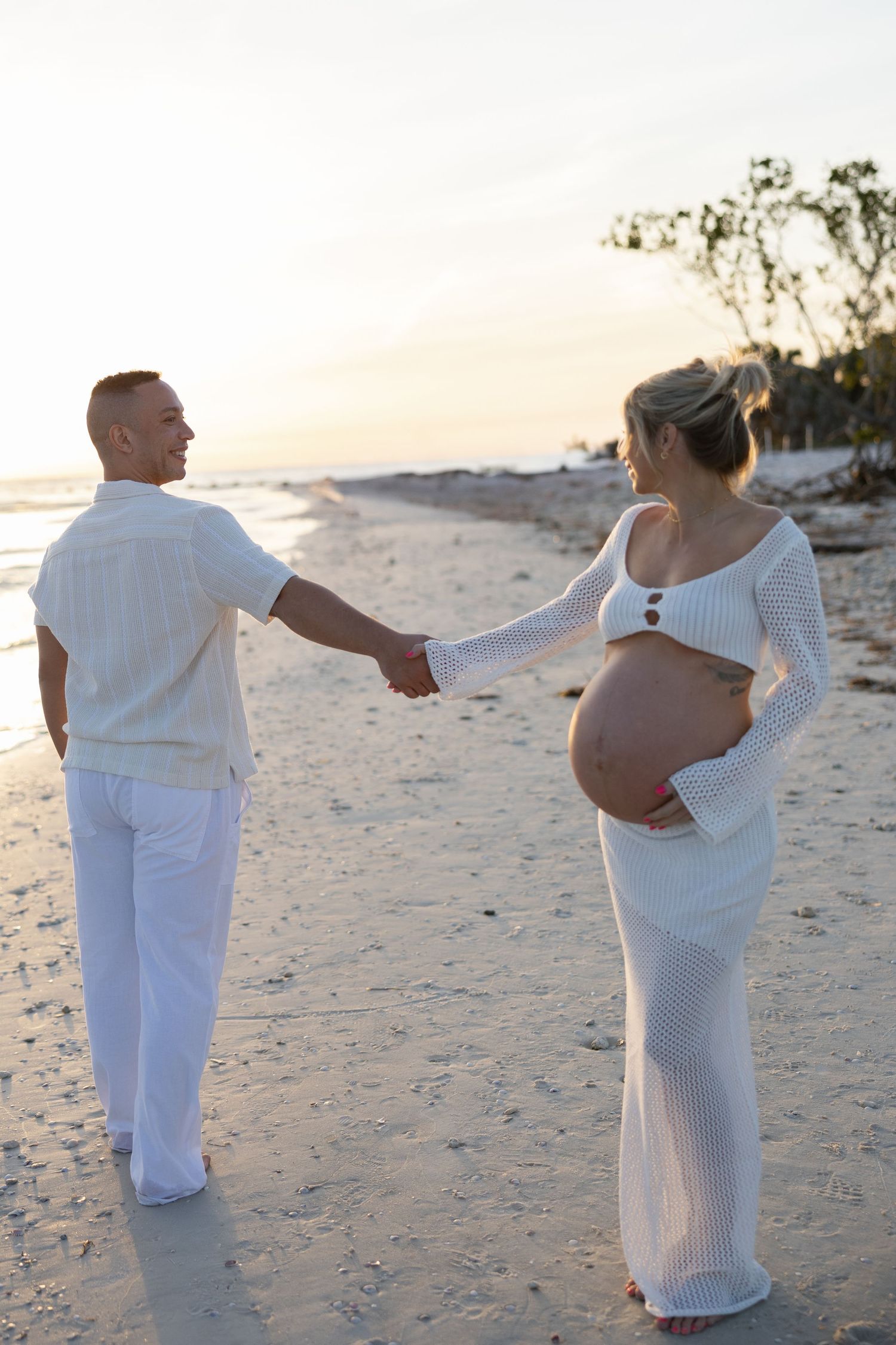 Pregnant couple in white outfits holding hands on beach during a beautiful sunset.
