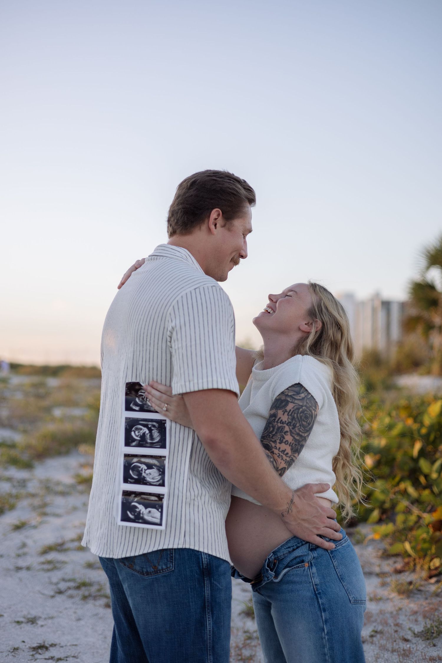 Expecting couple embracing on beach at sunset, holding ultrasound photos, woman laughing joyfully.