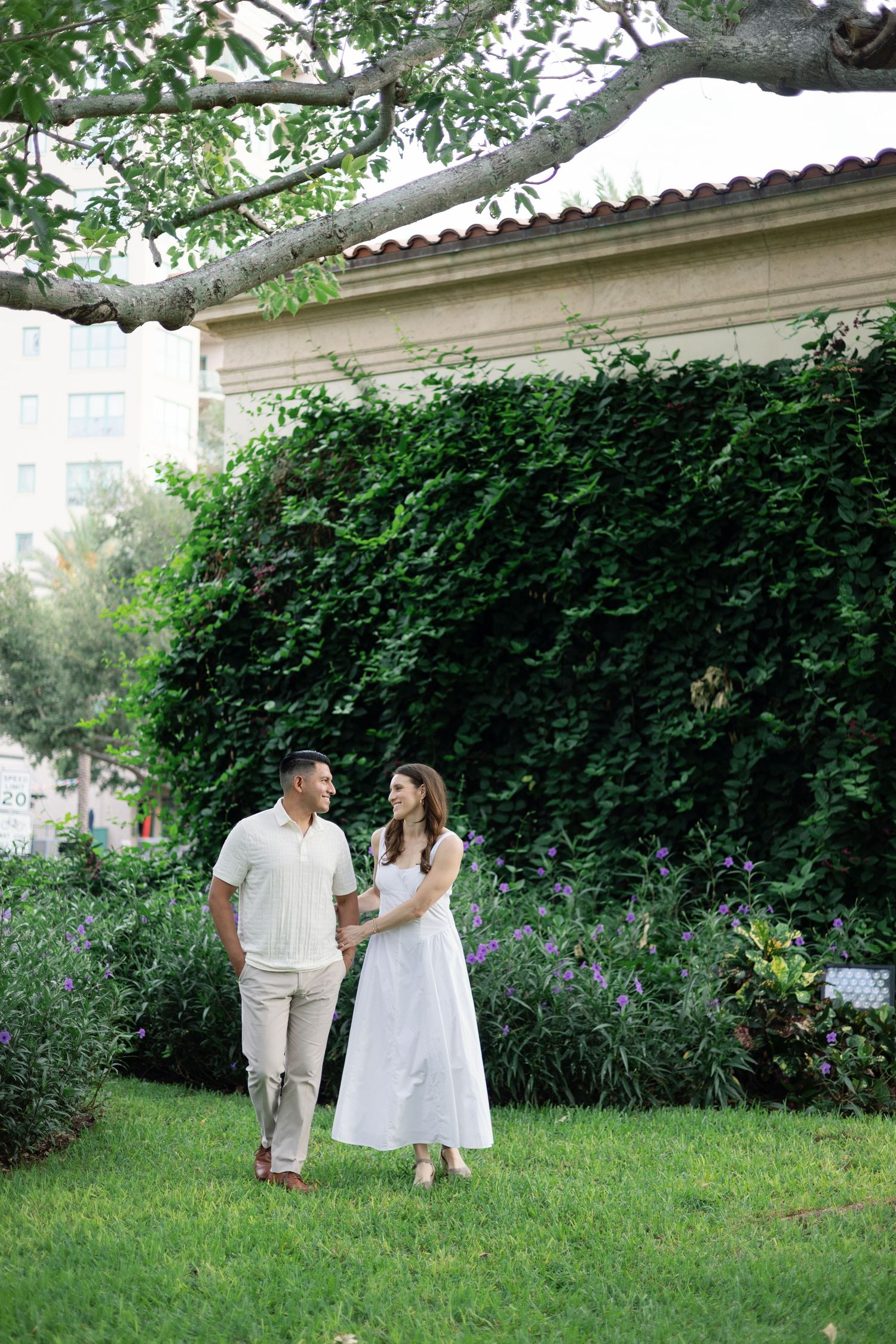 Couple in white outfits holding hands and walking on green lawn in front of ivy-covered garden wall.