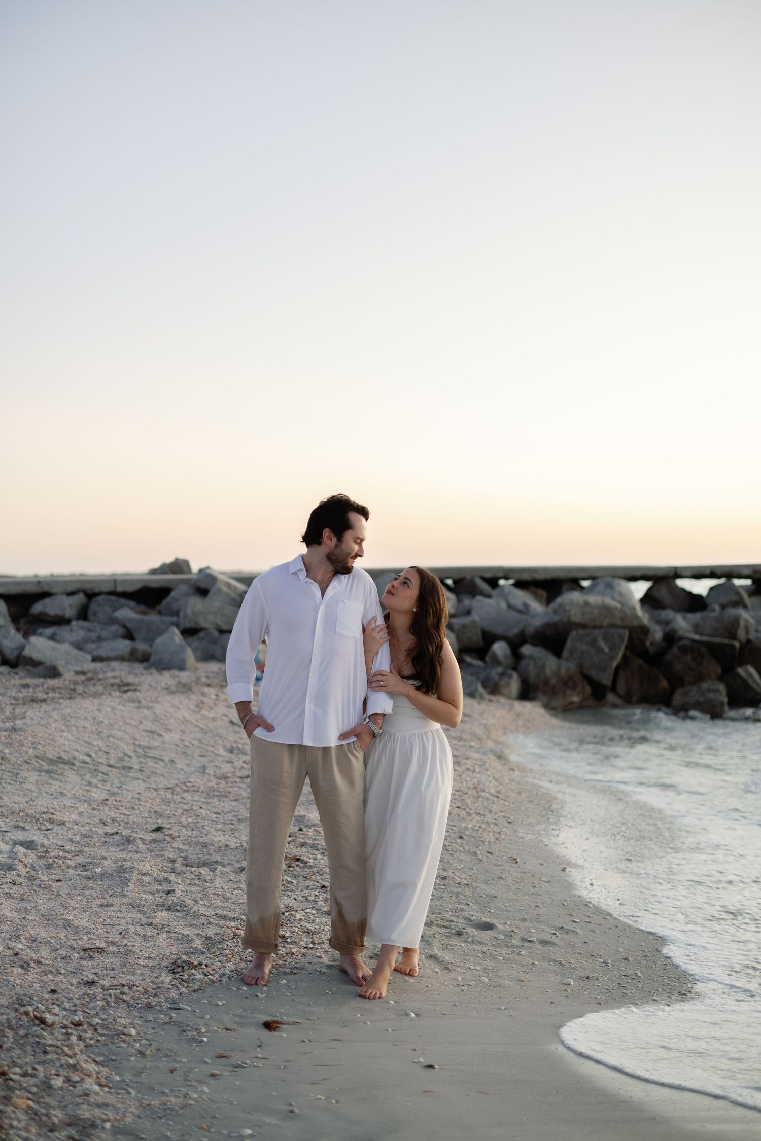 Couple walking barefoot on a serene beach at sunset, smiling at each other near rocky shoreline.
