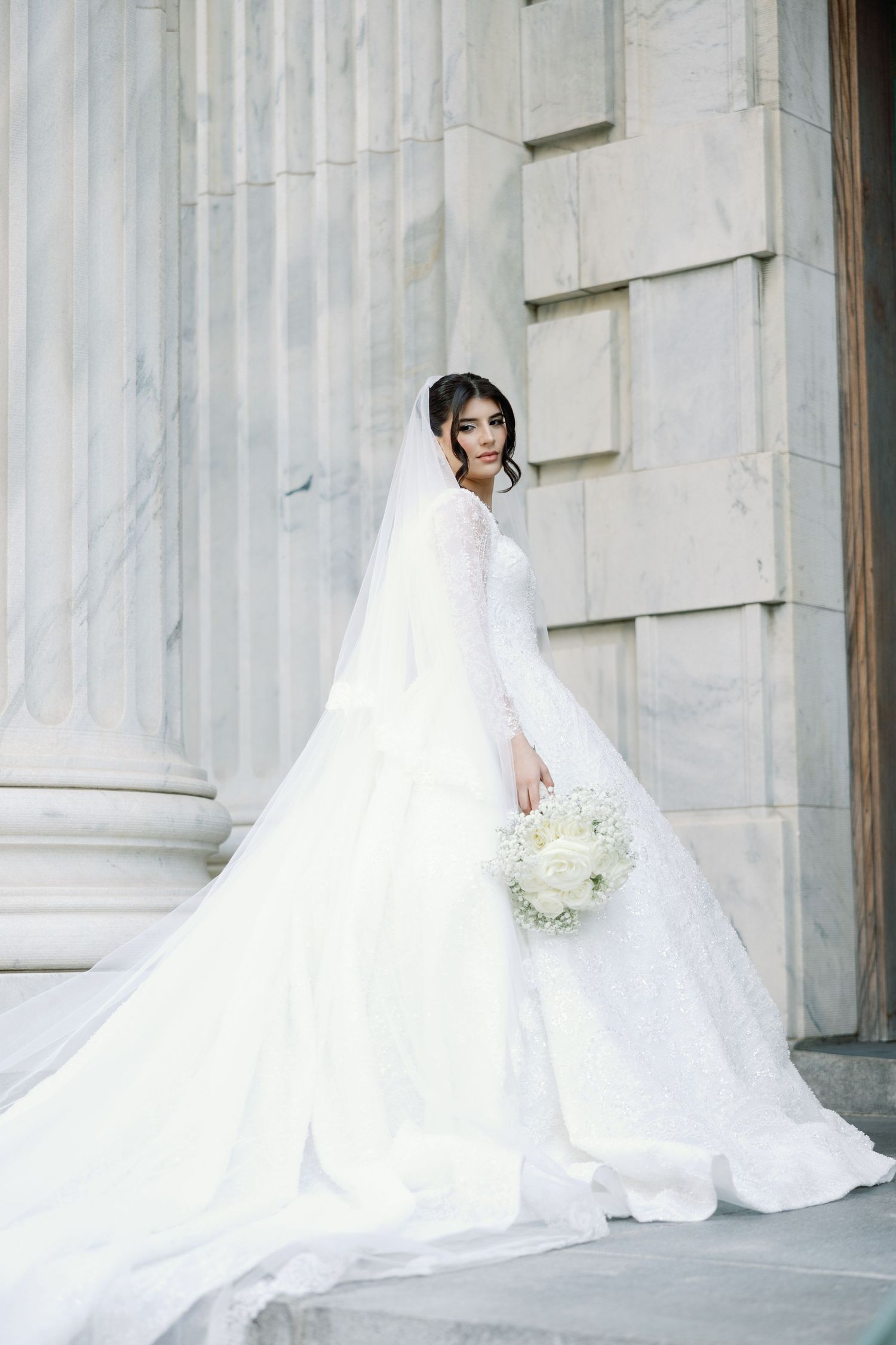 Elegant bride in long-sleeve lace ballgown holding white bouquet beside marble columns.