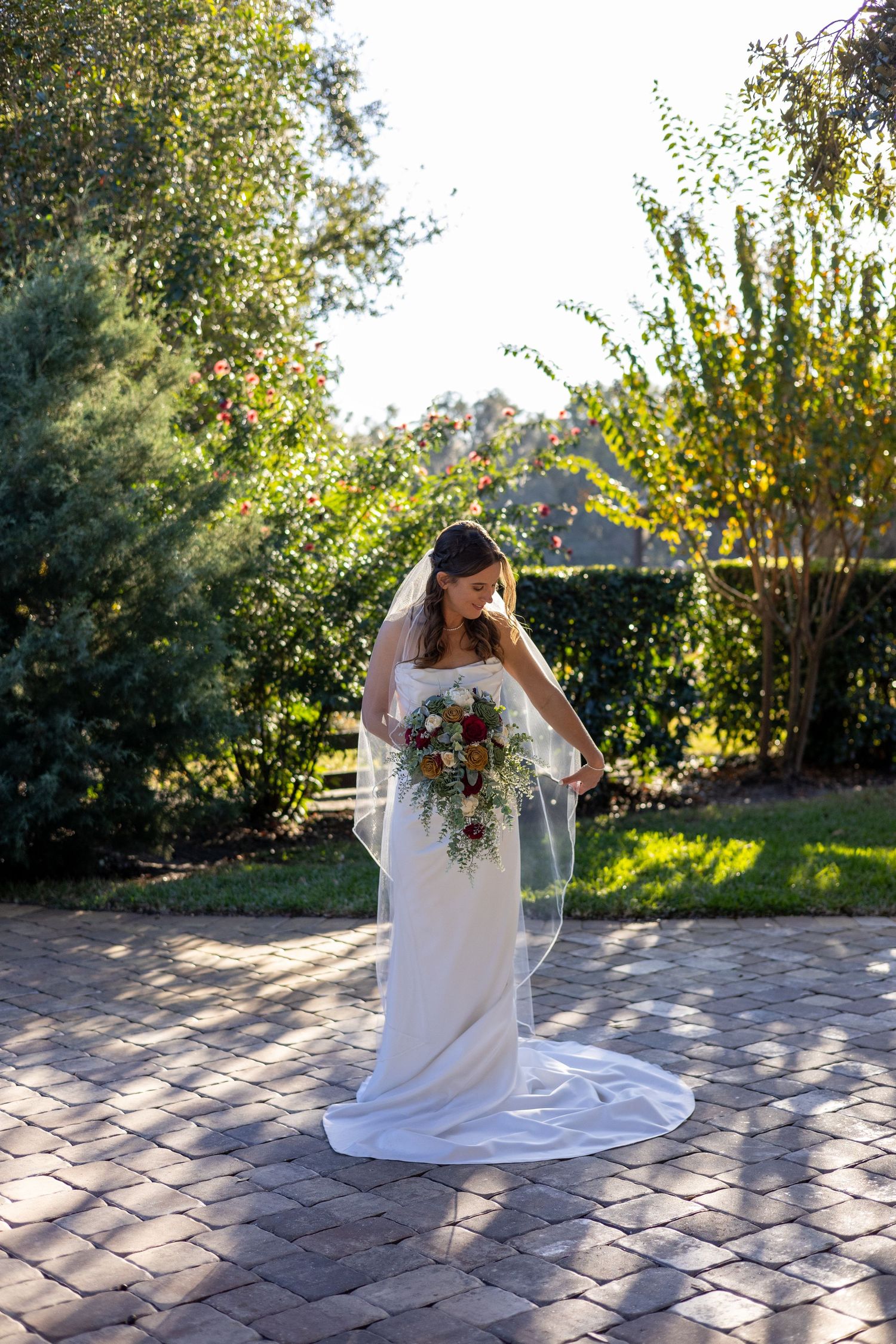 Bride in white gown and veil holding a floral bouquet on a brick patio in a sunlit garden.