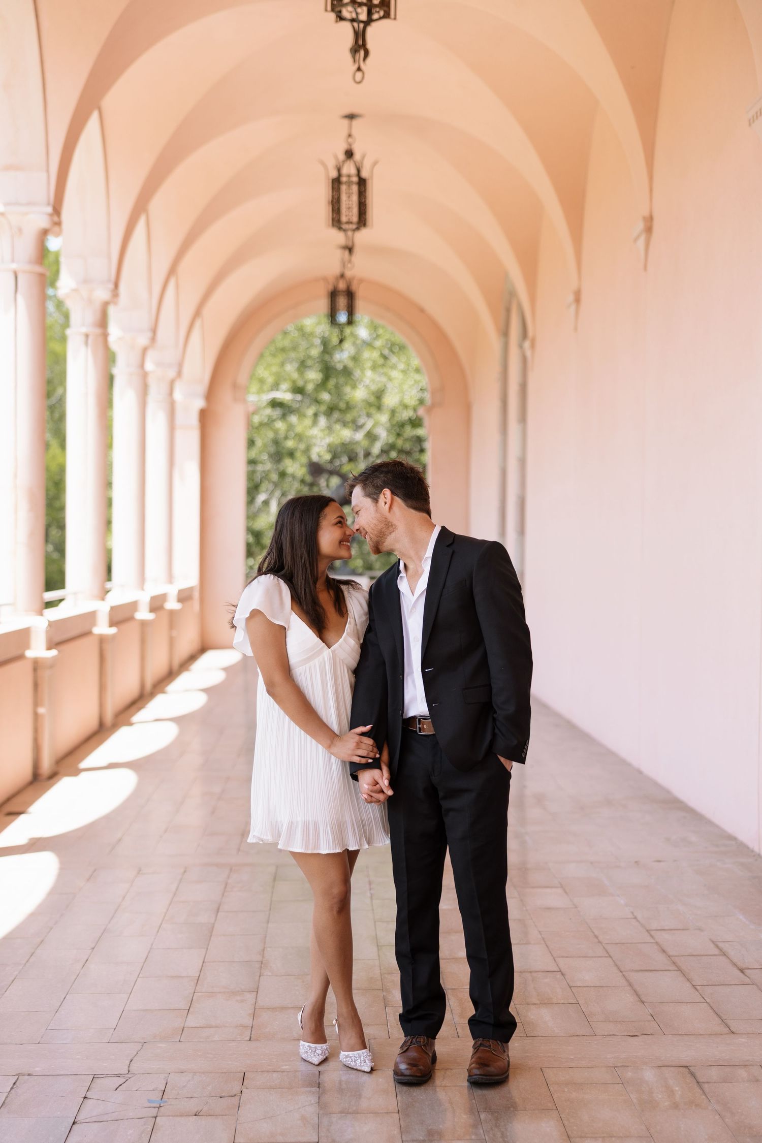 An elegant couple shares a romantic moment in a beautiful arched corridor of a pink historic building.