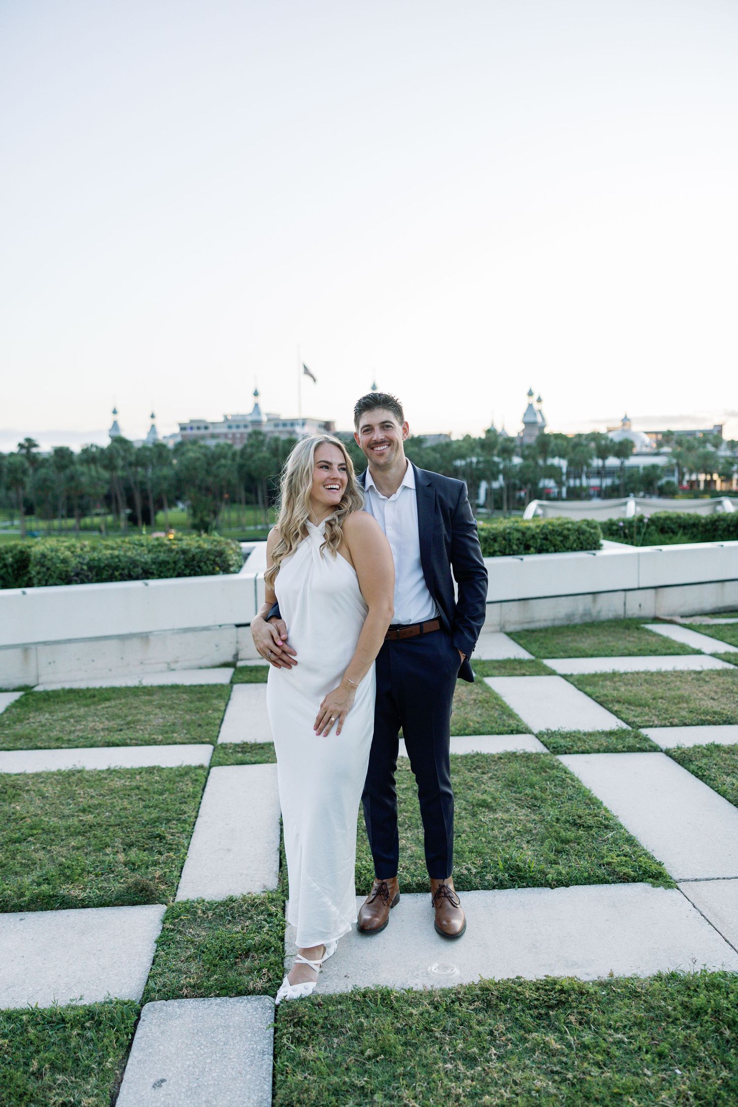 Happy couple in white dress and navy suit posing on checkered garden patio at dusk.