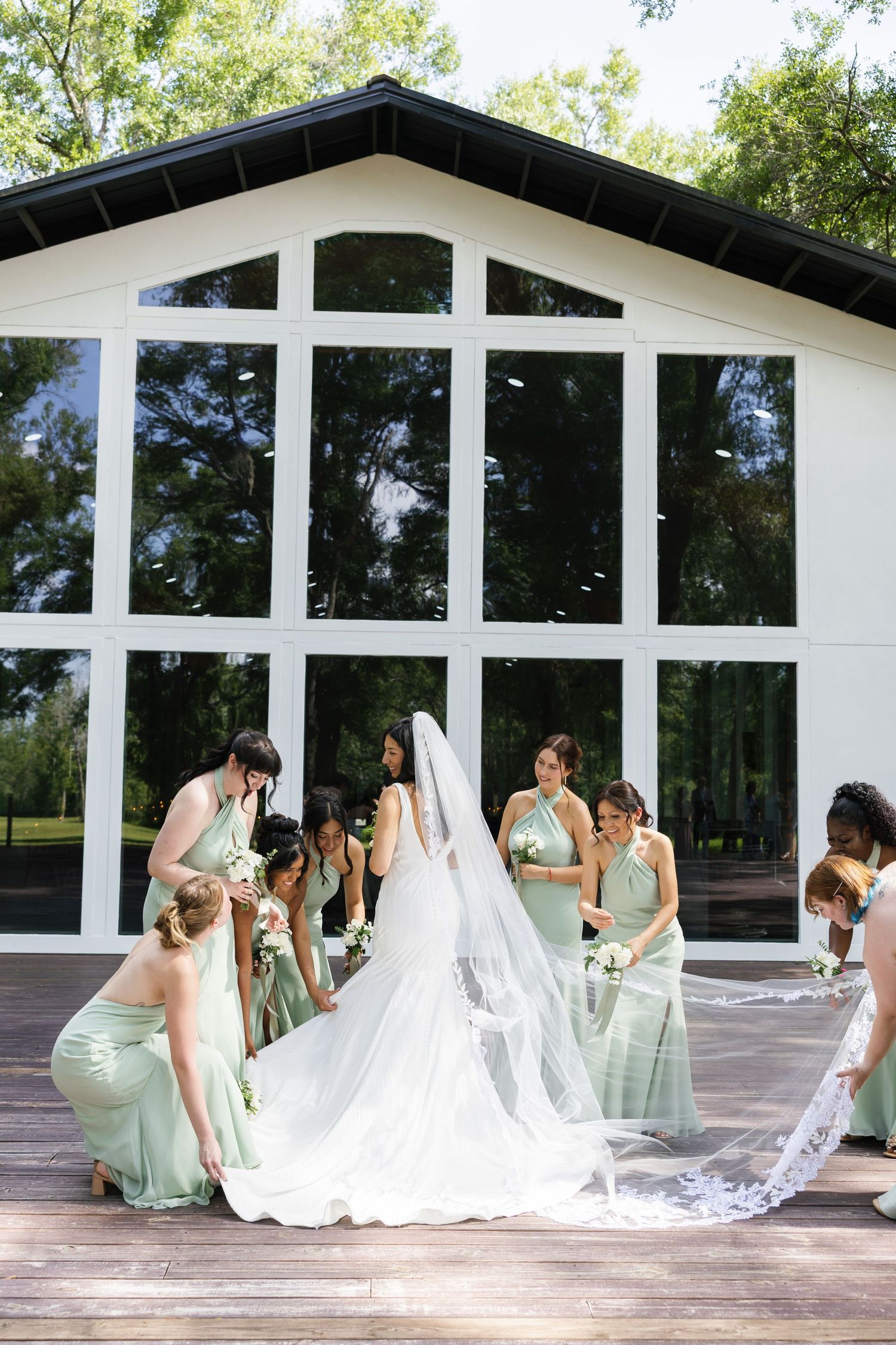 Bridesmaids in mint green dresses arrange bride's cathedral veil on wooden deck outside venue.
