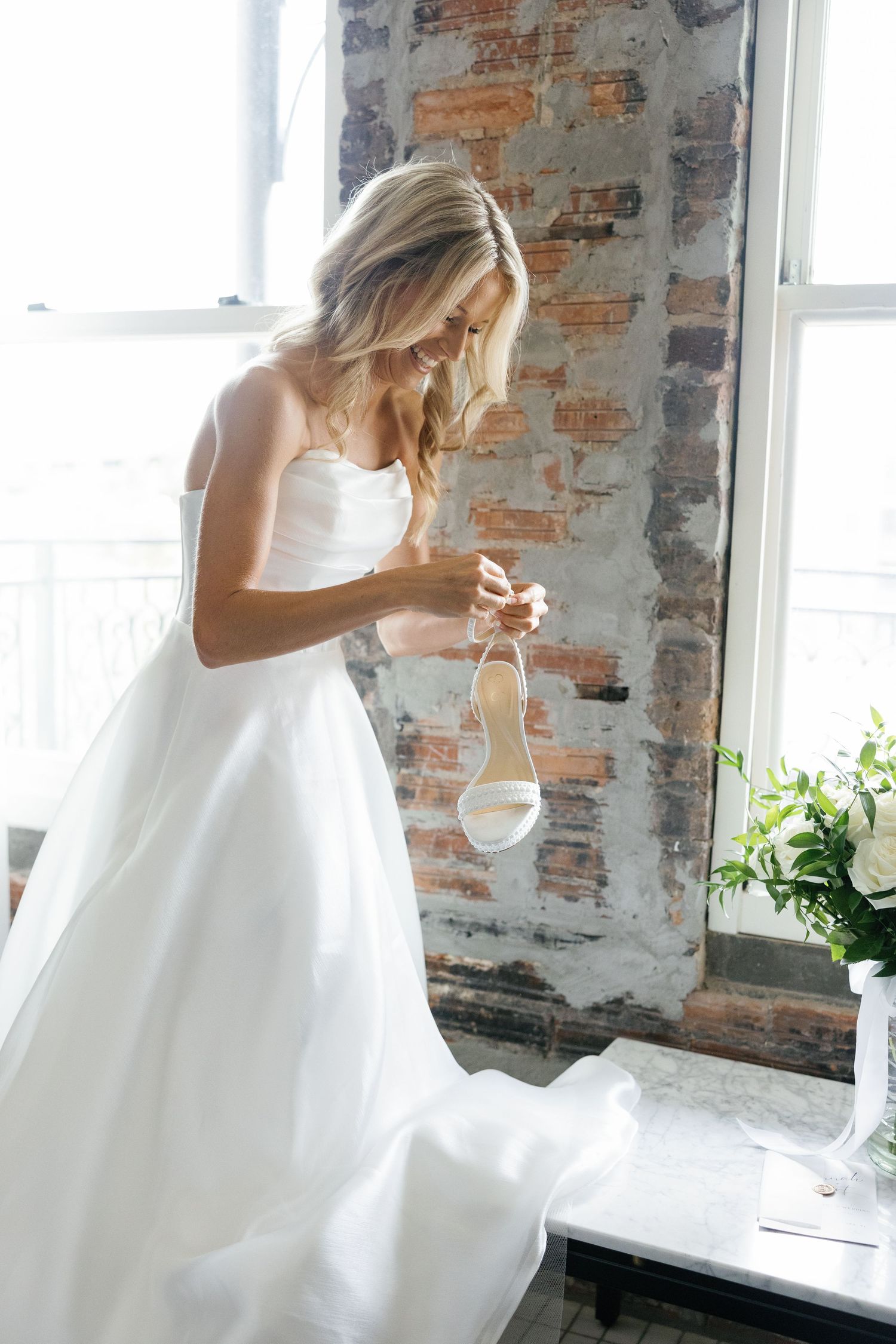 Bride in strapless white ballgown examines white heeled wedding shoe near sunlit exposed brick window.
