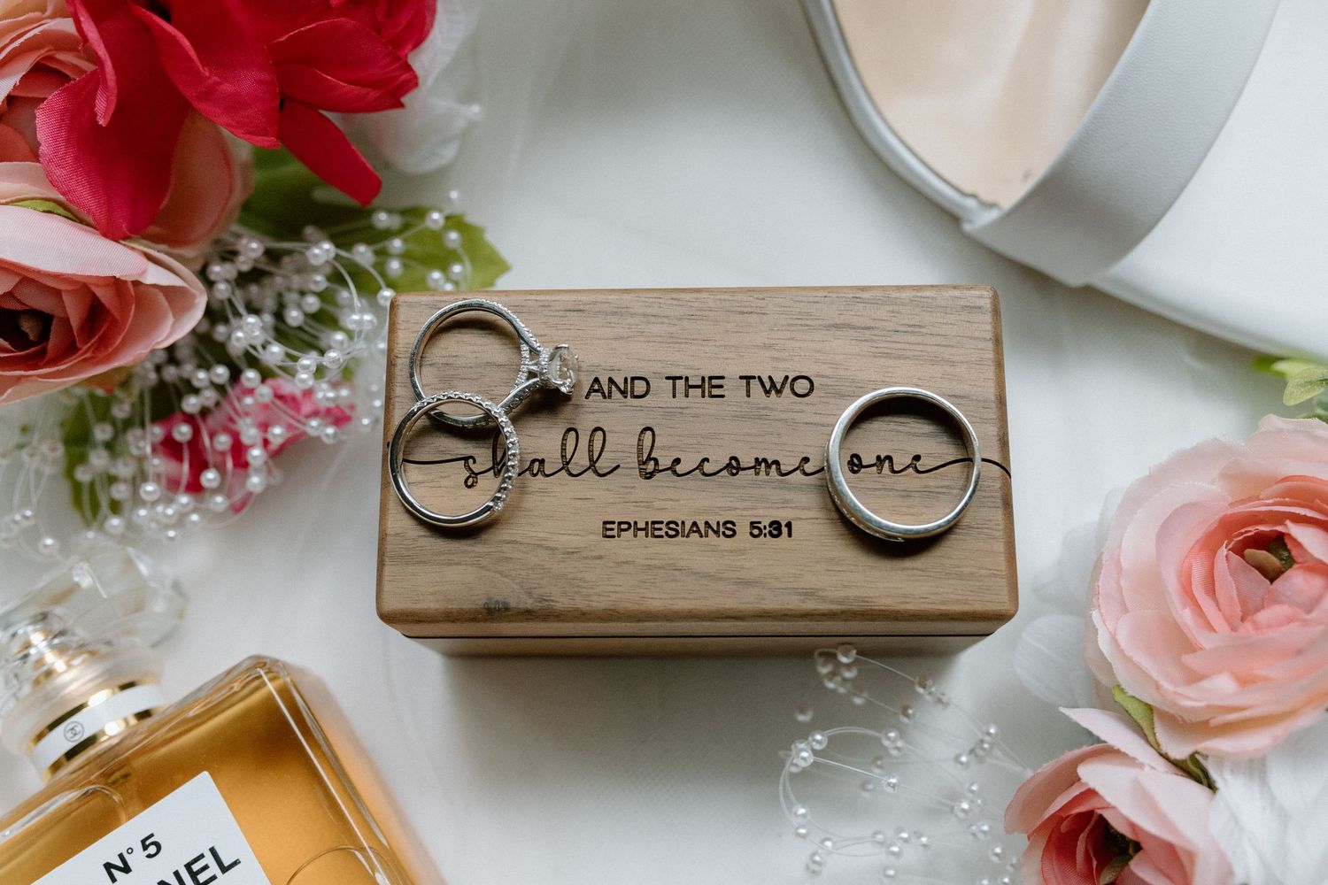 Wedding rings displayed on wooden box engraved with Ephesians 5:31 surrounded by pink flowers.