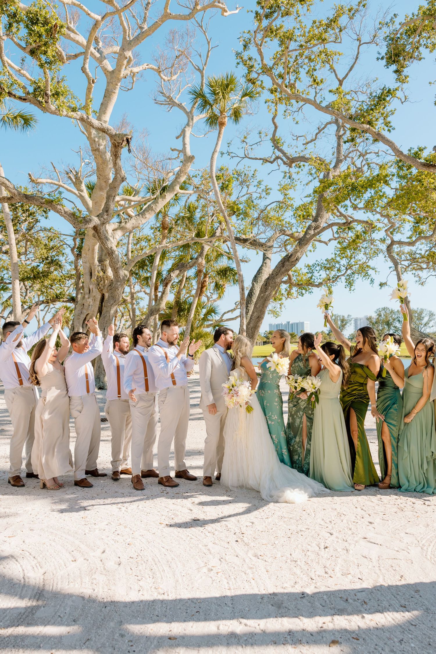 Wedding party celebrating on sandy beach, bridesmaids in green gowns and groomsmen in tan suits under oak trees.