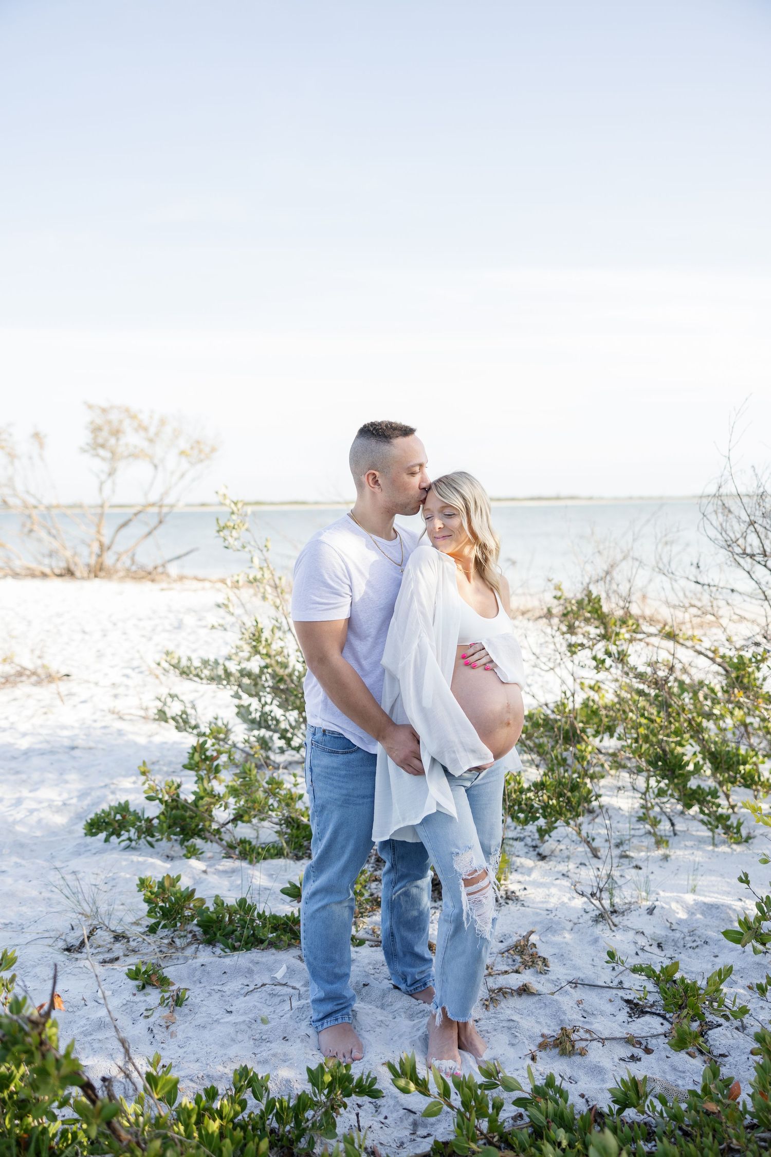 Couple embracing on white sand beach, man kissing pregnant woman's temple near coastal shrubs.