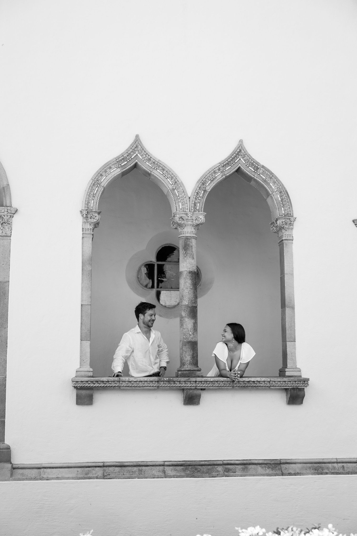A couple sits in ornate Gothic arched windows of a white historic building in a black and white photo.