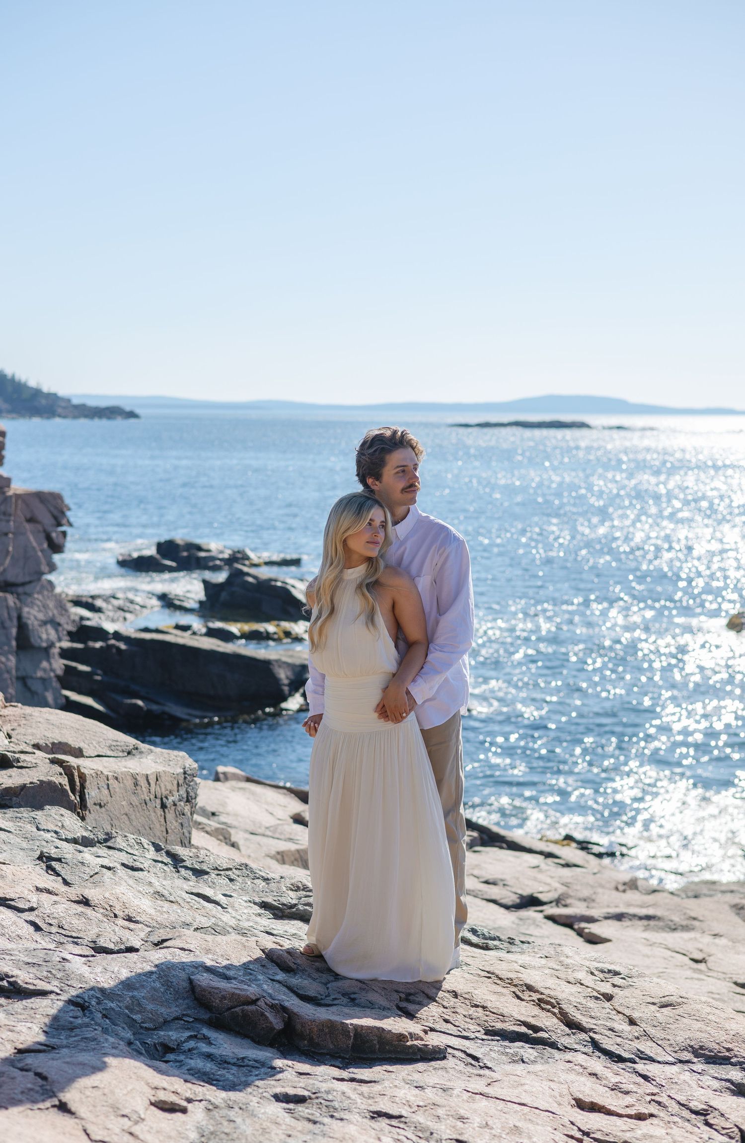 Couple embracing on rocky shoreline with sparkling blue ocean and clear sky behind them.