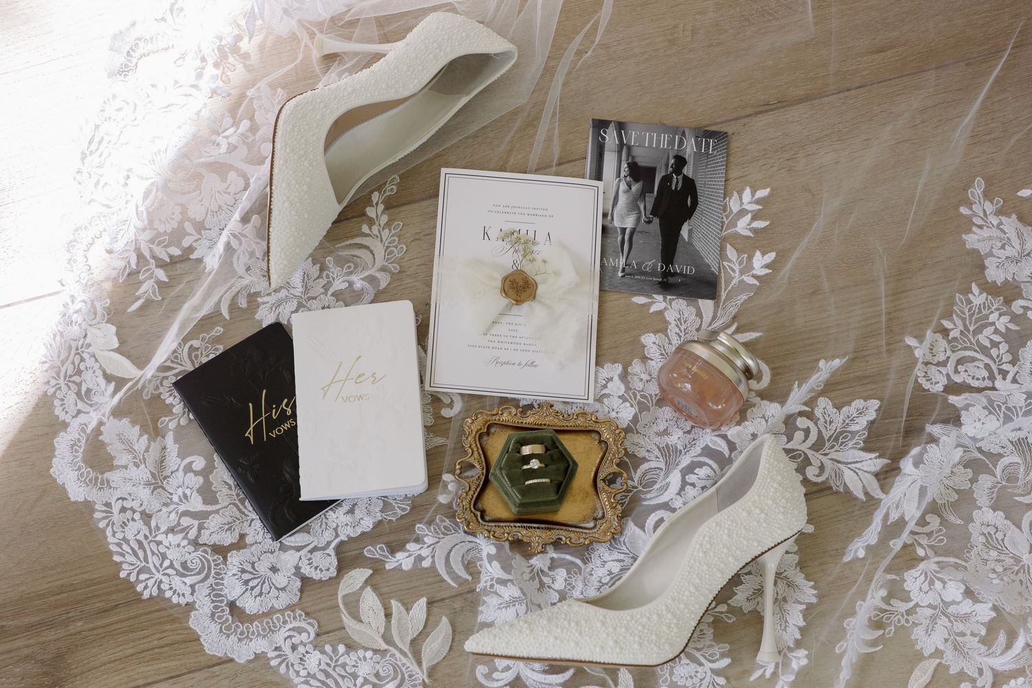 Flat lay of wedding details including lace veil, ivory heels, invitation suite, vintage brooch, and floral perfume bottle.