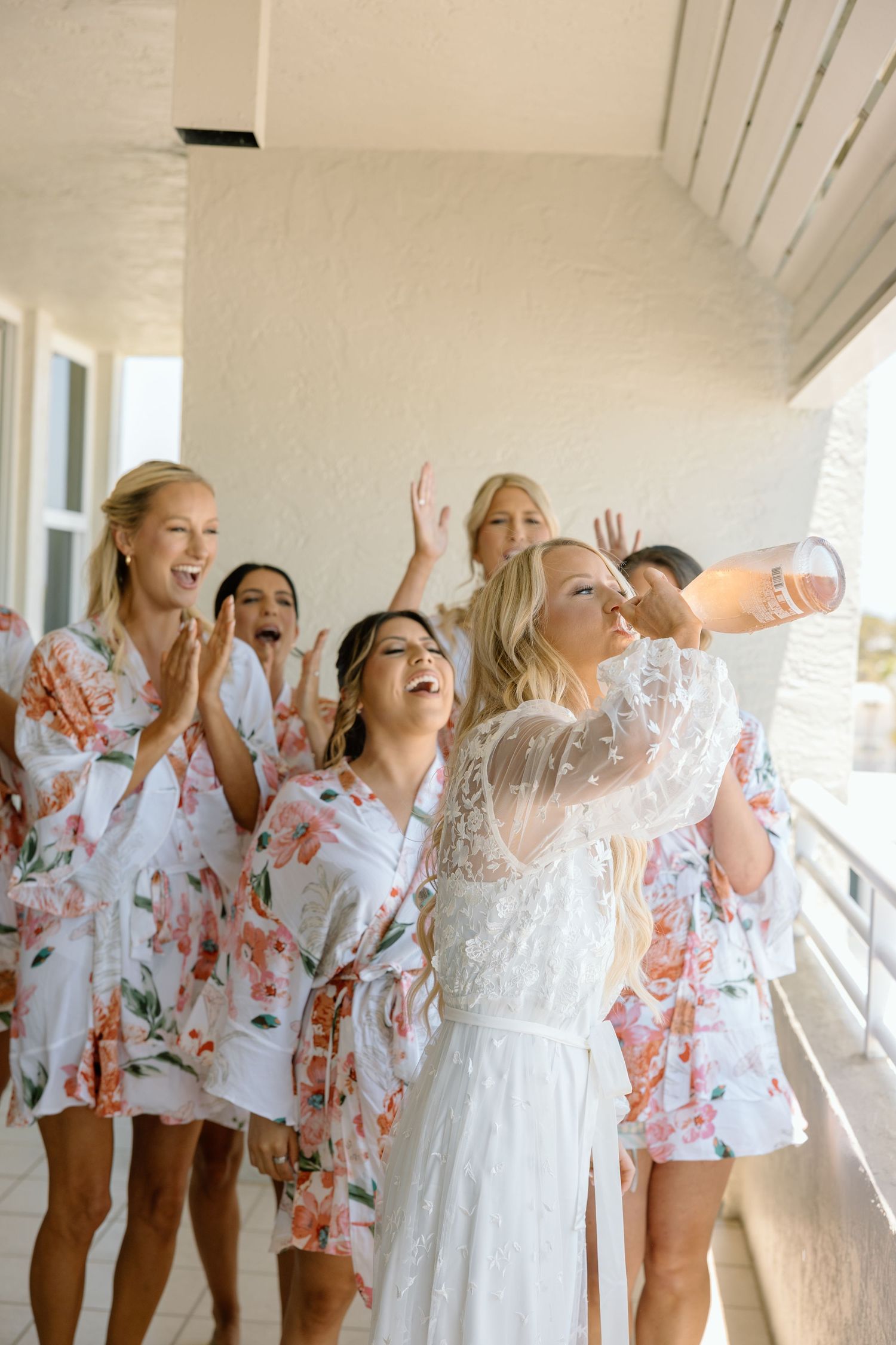 Excited bride and bridesmaids in floral robes celebrating on balcony during wedding morning preparations.